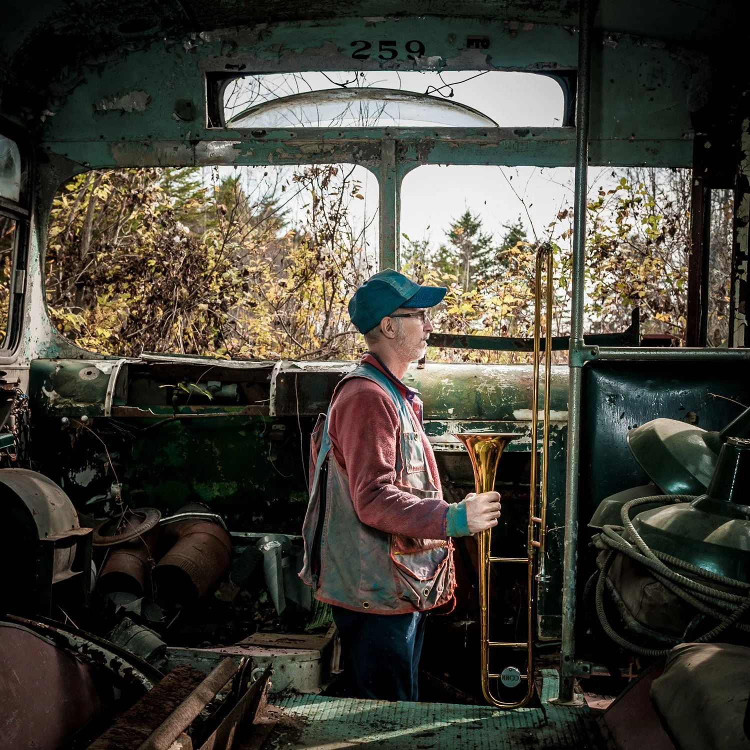 Un homme portant une casquette, une veste multicolore et des lunettes, tenant un trombone, se tient à l'intérieur d'une carcasse de bus abandonnée, avec une végétation automnale visible par la fenêtre.