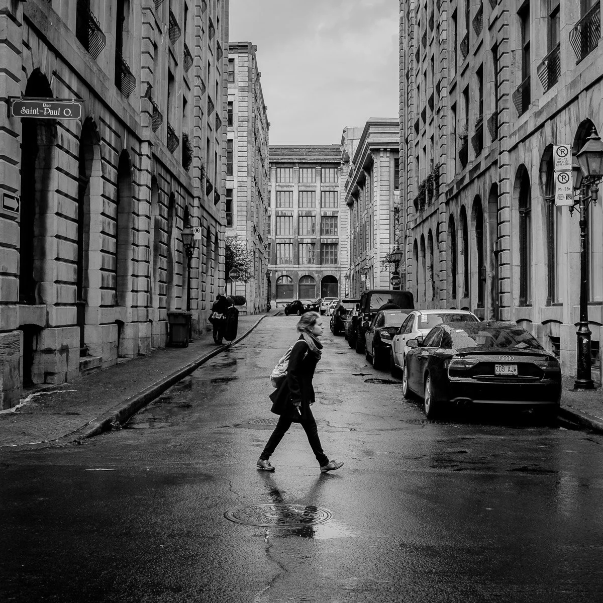 Une rue urbaine en noir et blanc avec des bâtiments en pierre, des voitures stationnées le long du trottoir et une femme marchant sur la chaussée mouillée.