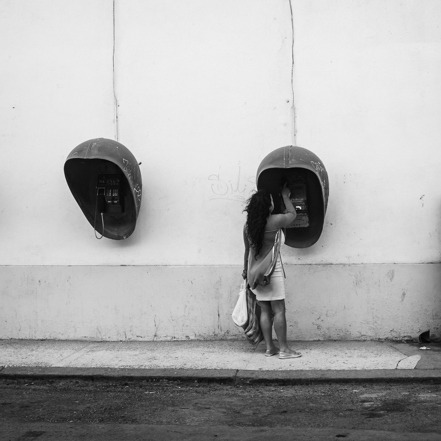 Une femme avec de longues cheveux bouclés, portant un t-shirt et une jupe, utilise un téléphone public fixé à un mur blanc. Elle a un sac et une serviette en tissu. La scène est en noir et blanc et se déroule au bord de la rue.