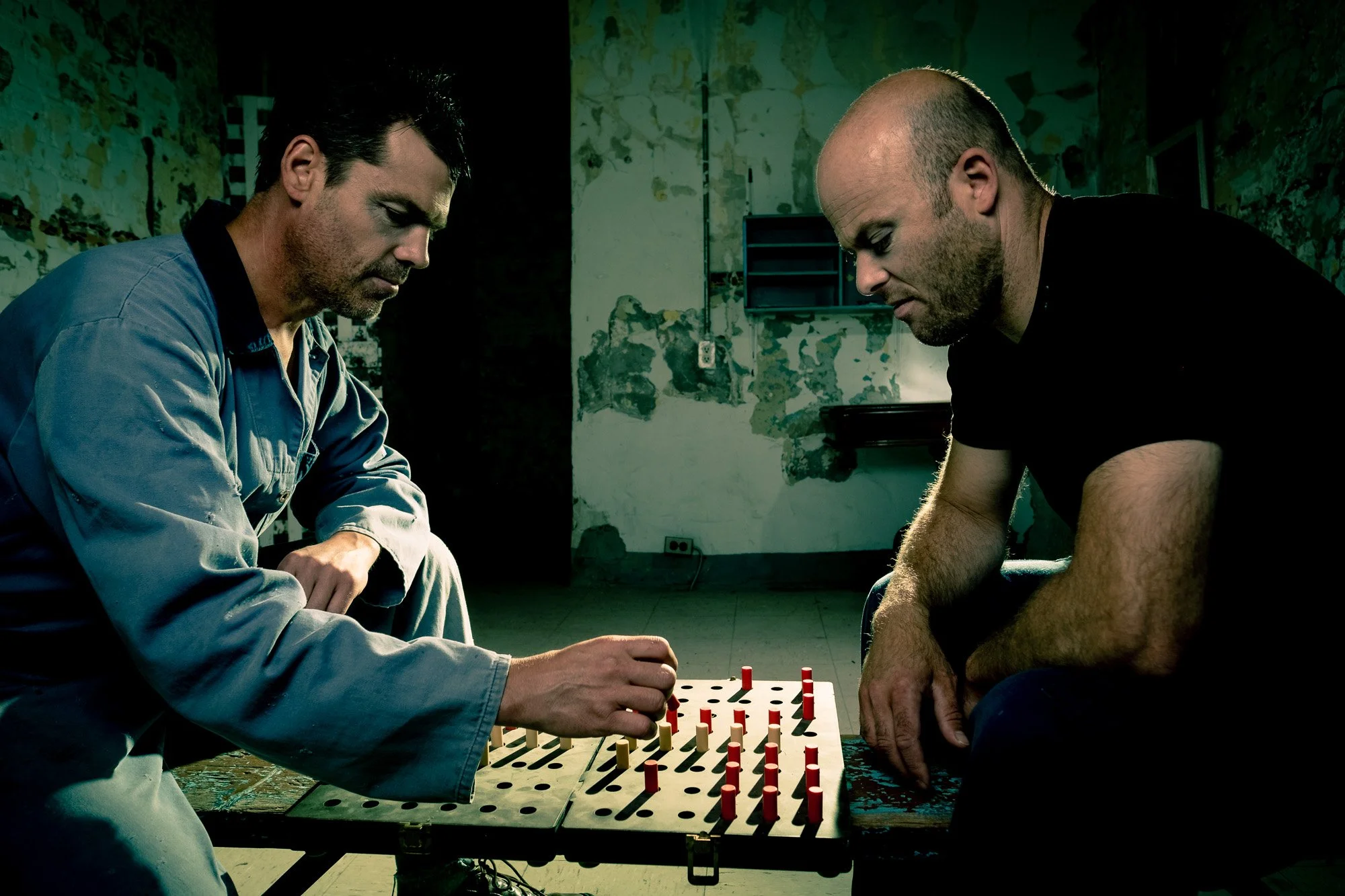 Deux hommes jouent à un jeu de société, probablement aux échecs ou jeu de stratégie, dans une pièce aux murs usés et dégradés, avec une atmosphère sombre.