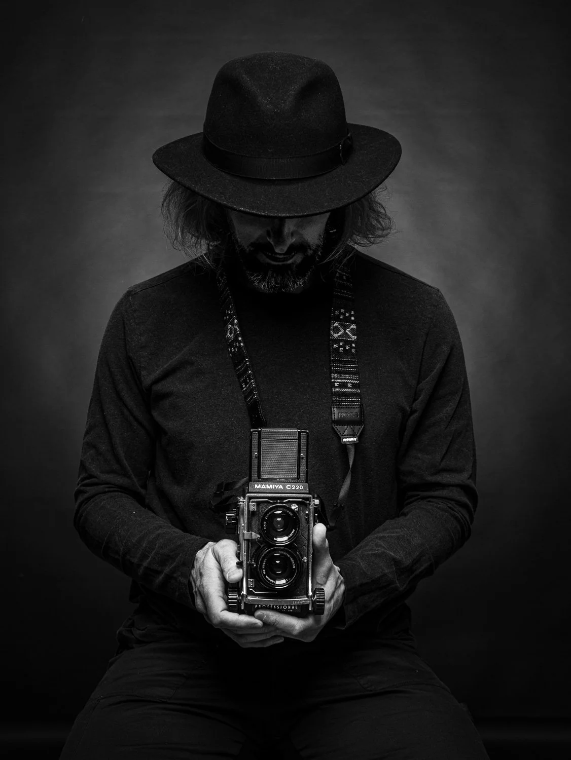 Homme portant un chapeau noir et tenant un vieil appareil photo réflex numérique, en vêtements noirs, vu de face, dans un studio noir et blanc.