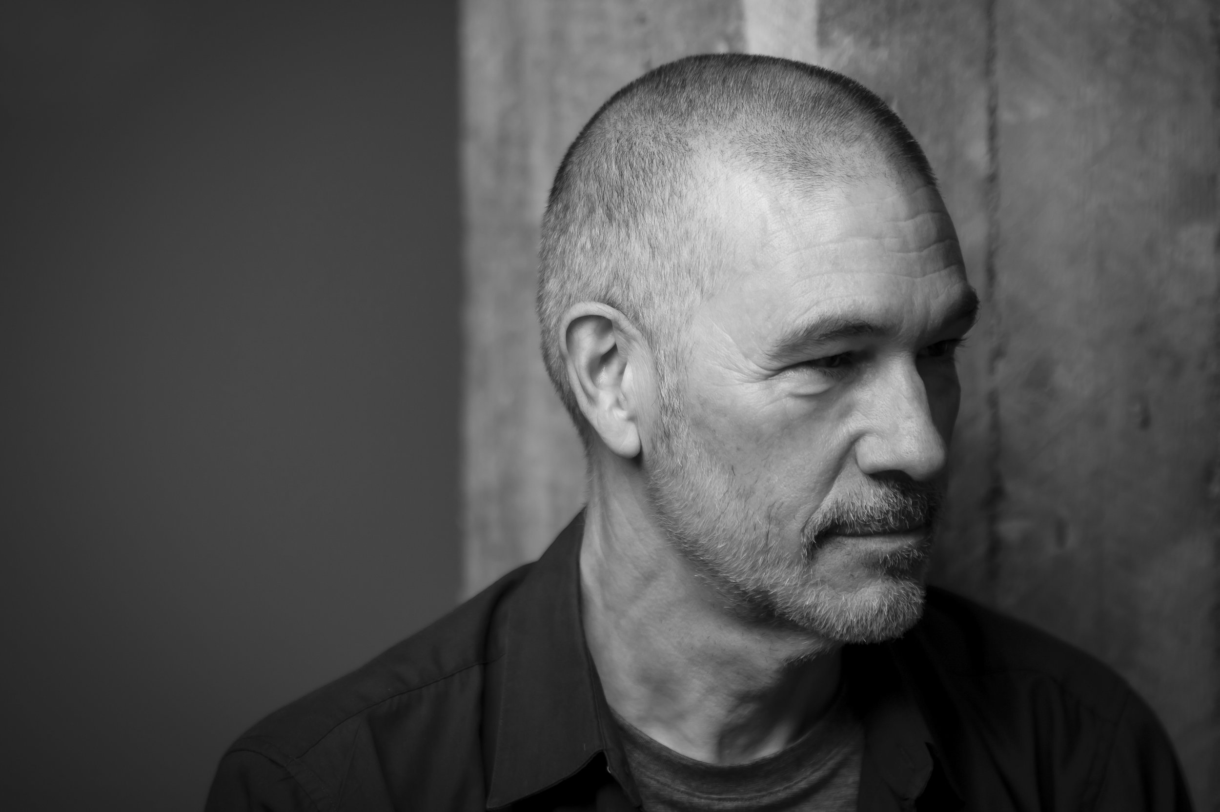 Portrait en noir et blanc d'un homme barbu et cheveux courts, regardant vers la droite, avec un mur en béton en arrière-plan.