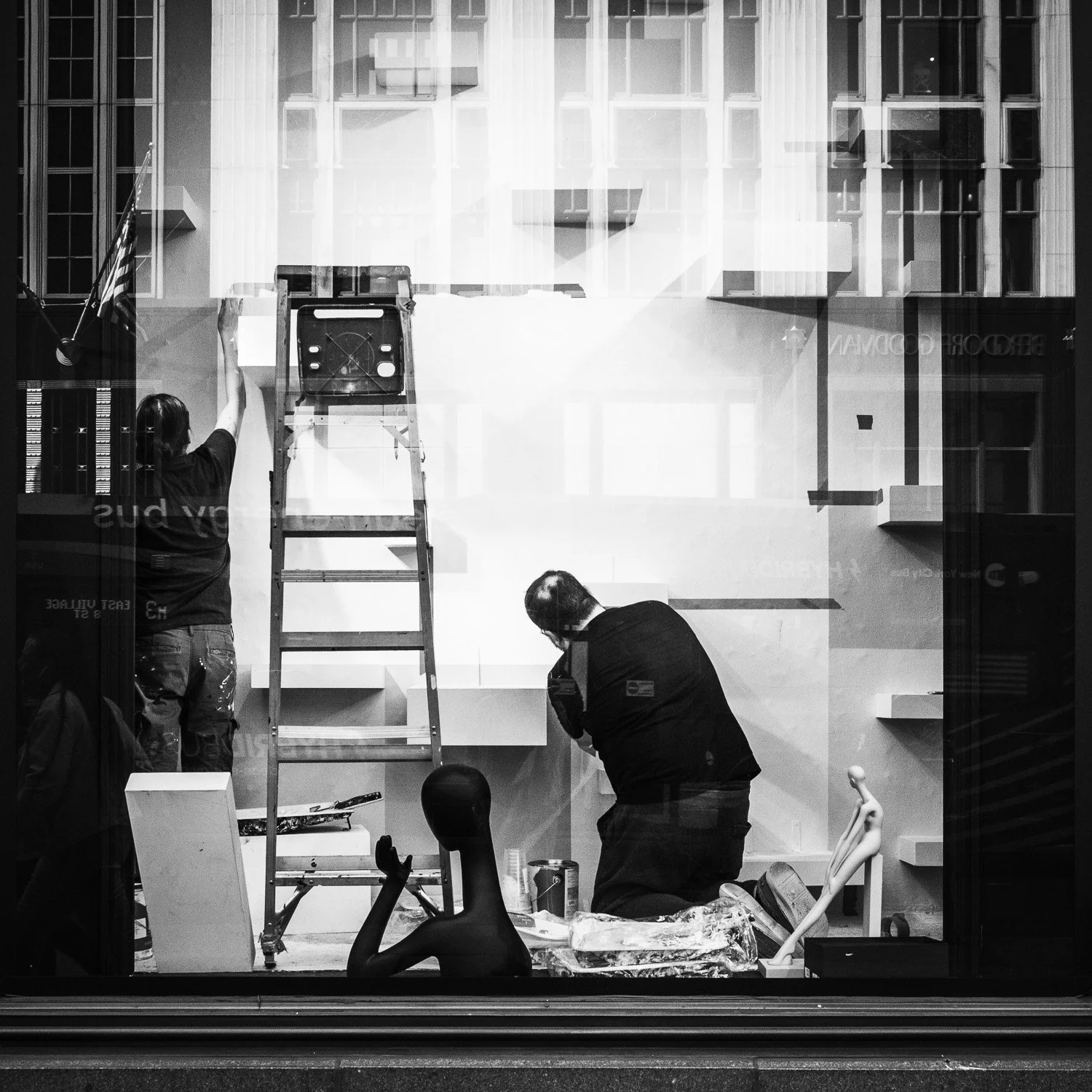 Vue de l'extérieur d'un magasin, avec deux personnes à l'intérieur. L'une de elles est debout près d'une échelle avec un drapeau américain. L'autre est penchée en avant, semblant travailler sur un projet ou une installation d'une vitrine.