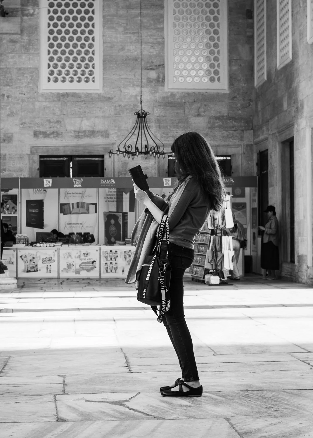 Une femme debout, regardant son téléphone, dans un espace intérieur avec un plafond haut, un chandelier suspendu, et des stands de livres ou de marchandises en arrière-plan, en noir et blanc.