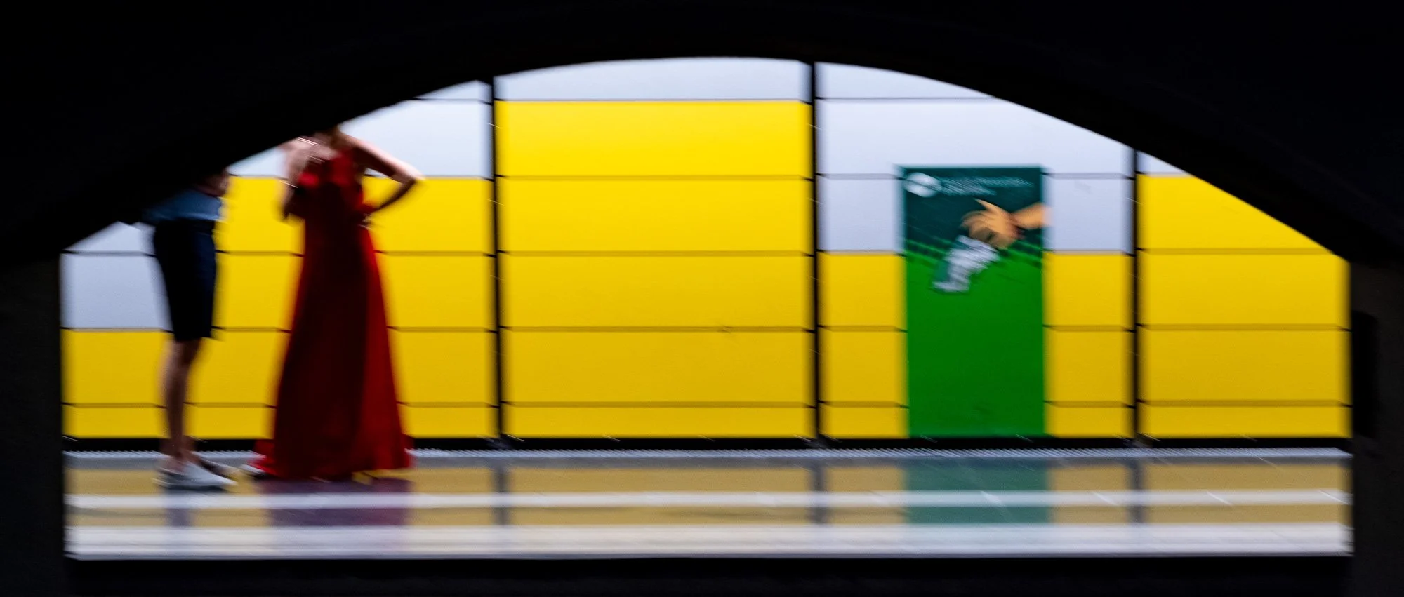 Une photo floue d'une ouverture dans un tunnel montrant deux femmes. L'une porte une robe rouge et les talons, l'autre un haut bleu et un jean court blanc, avec un sac. À l'arrière-plan, un mur avec des blocs de couleurs jaune et vert, dont un avec u