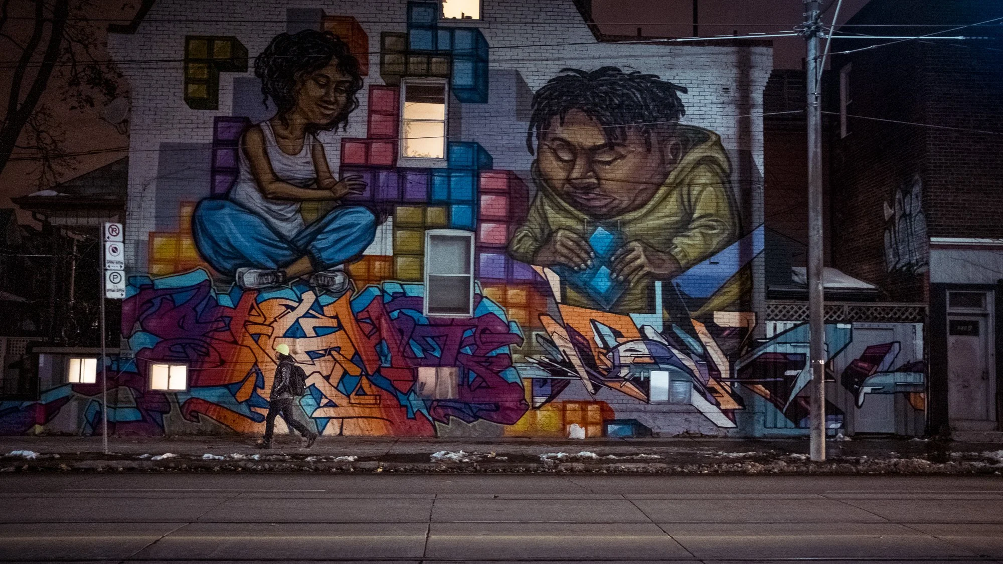 Une fresque murale représentant deux enfants, l'un jouant avec des blocs colorés et l'autre utilisant une tablette, avec des graffitis en bas. La scène est en plein air, en nocturne.