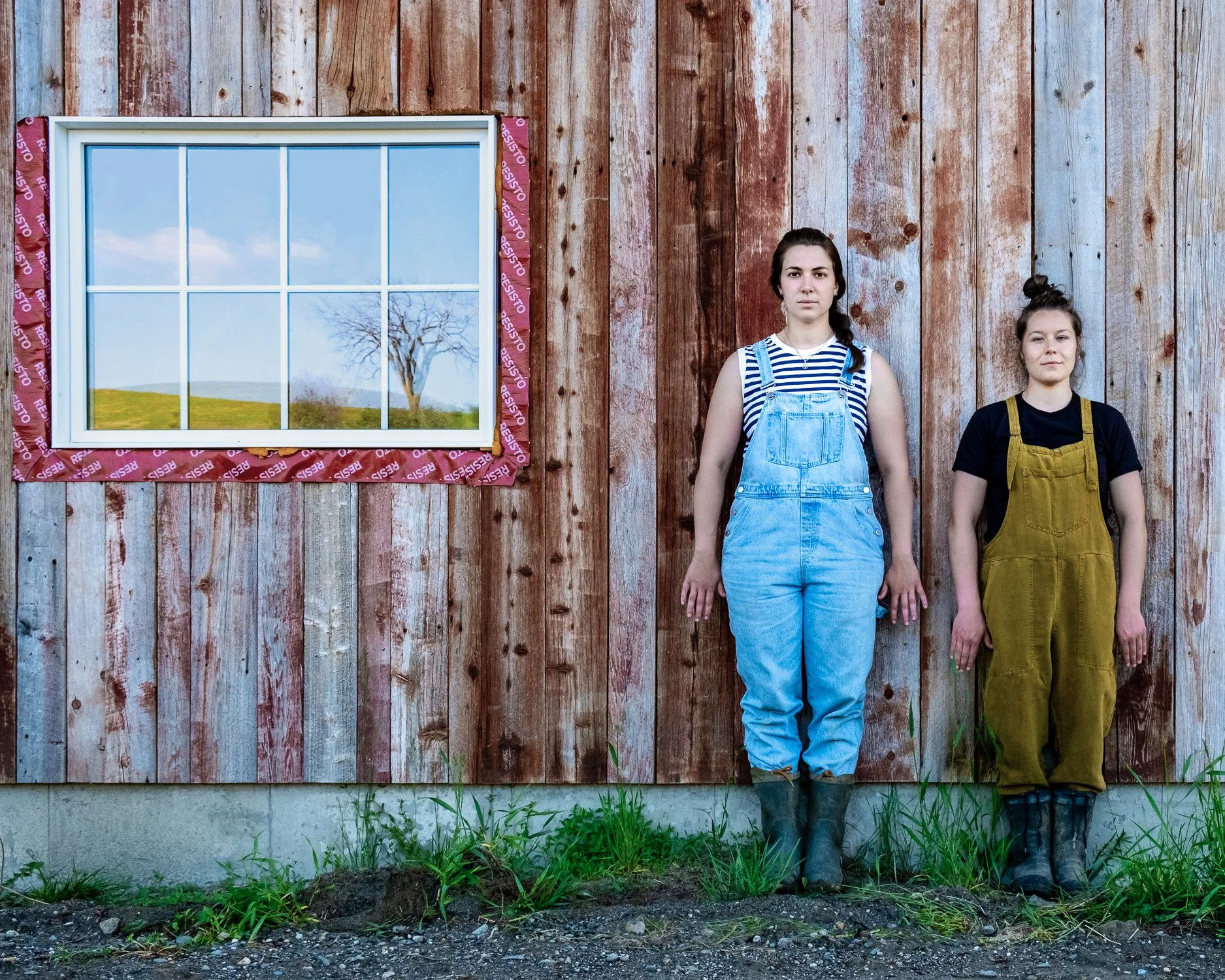 Deux jeunes femmes se tiennent debout contre un mur en bois. L'une porte une salopette en jean bleu, l'autre une salopette en coton kaki. Il y a une fenêtre à côté d'elles, qui reflète un arbre et un ciel bleu avec quelques nuages.