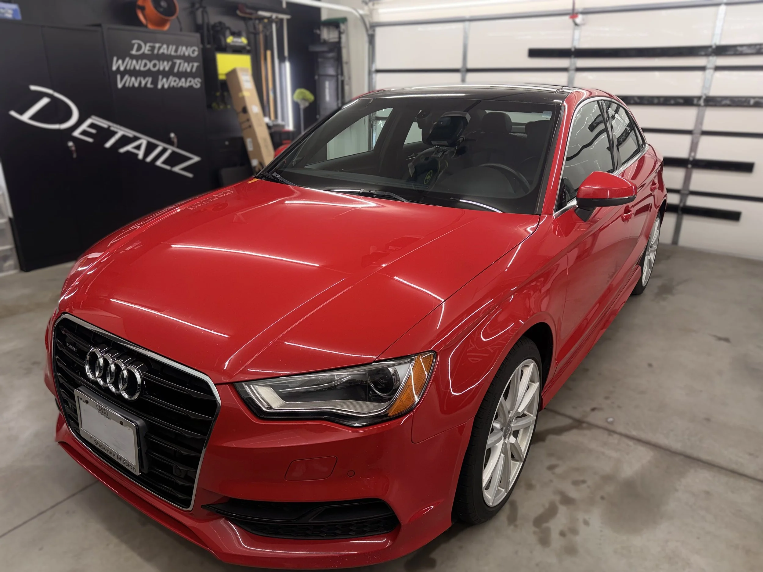 Red Audi sedan parked in a garage with a detailing and vinyl wrap station in the background.