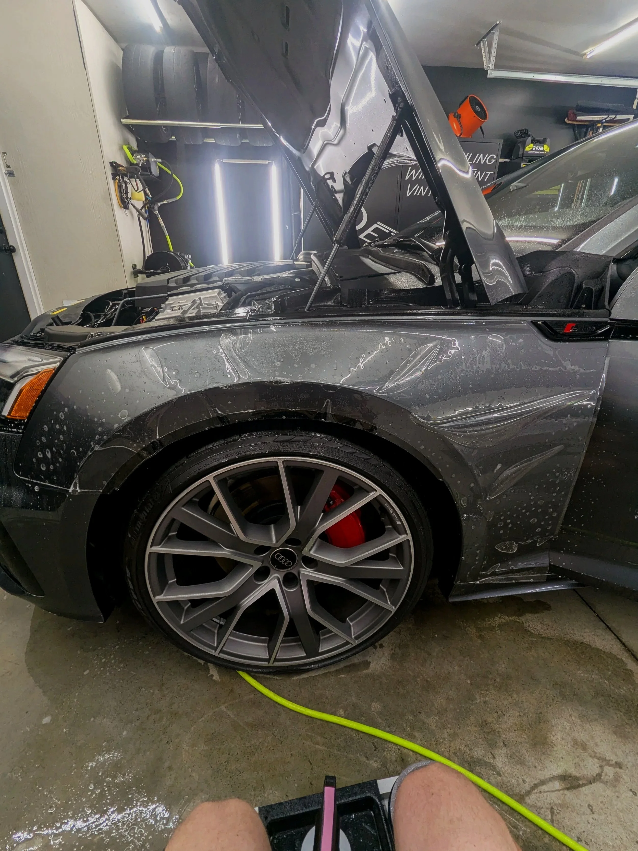 A black car with its hood open in a garage, showing exposed engine parts. The car's front left tire, with a red brake caliper, is visible. There are water spots on the car and on the garage floor, and a person is sitting nearby with legs visible. A green hose is on the floor, and garage tools and shelves are seen in the background.