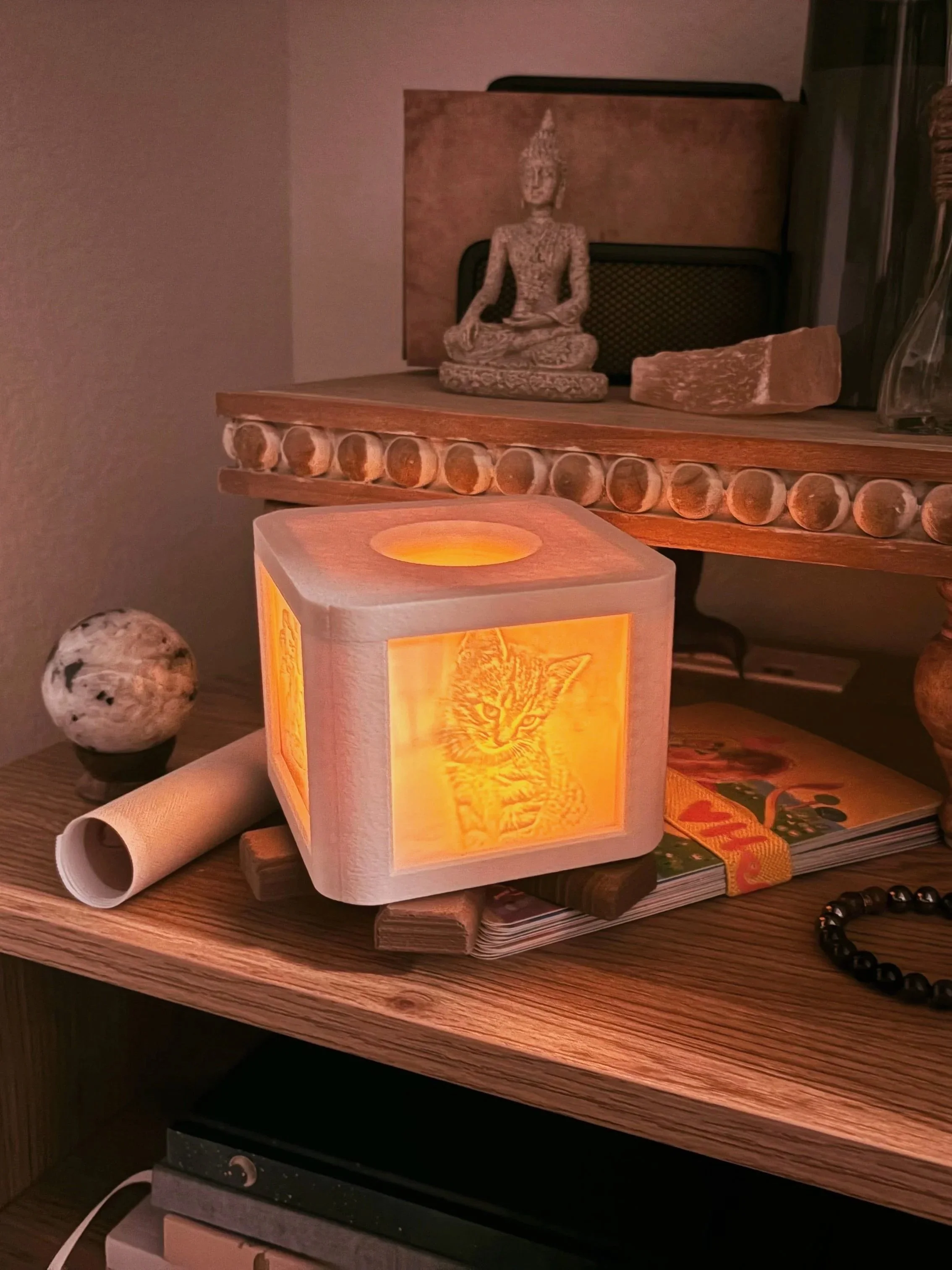 A small illuminated cube with a cat image on one side sits on a wooden surface, surrounded by various objects including a globe, rolled-up paper, a magazine, a beaded bracelet, and decorative statues in the background.