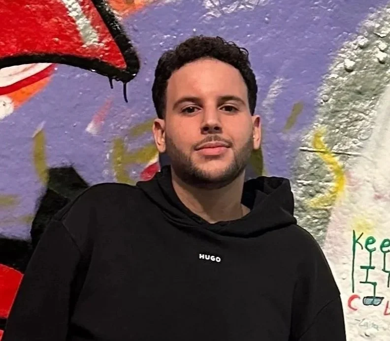 A young man with short, curly dark hair and a well-groomed beard, sitting at a restaurant or bar during the evening. He is wearing a black shirt with a plaid inner collar, a wristwatch, and is resting his chin on his hand. Behind him are warm lighting fixtures, other people in the background, and a window showing city lights and buildings outside.