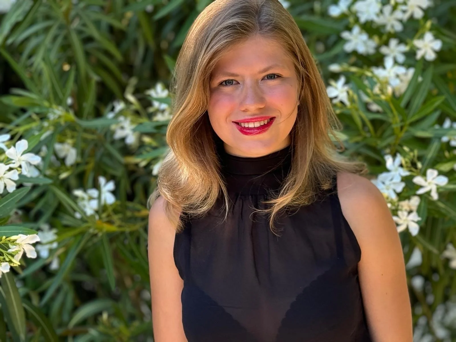 Young man in a graduation gown and tie standing in front of pink flowering bushes.