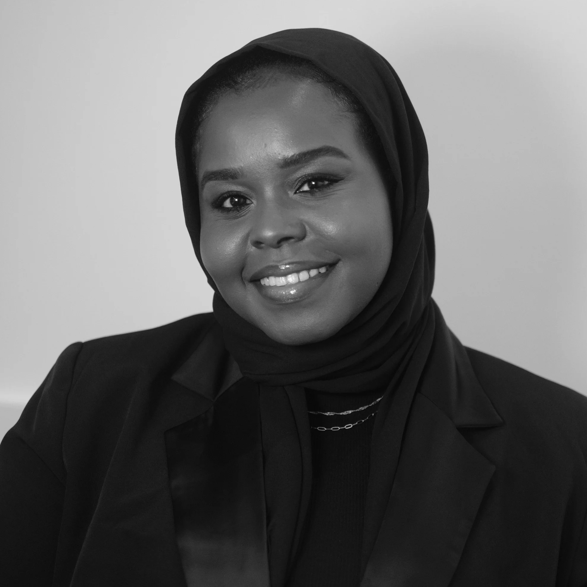 A woman wearing a headscarf and blazer, smiling and holding the lapels of her blazer, in black and white portrait.