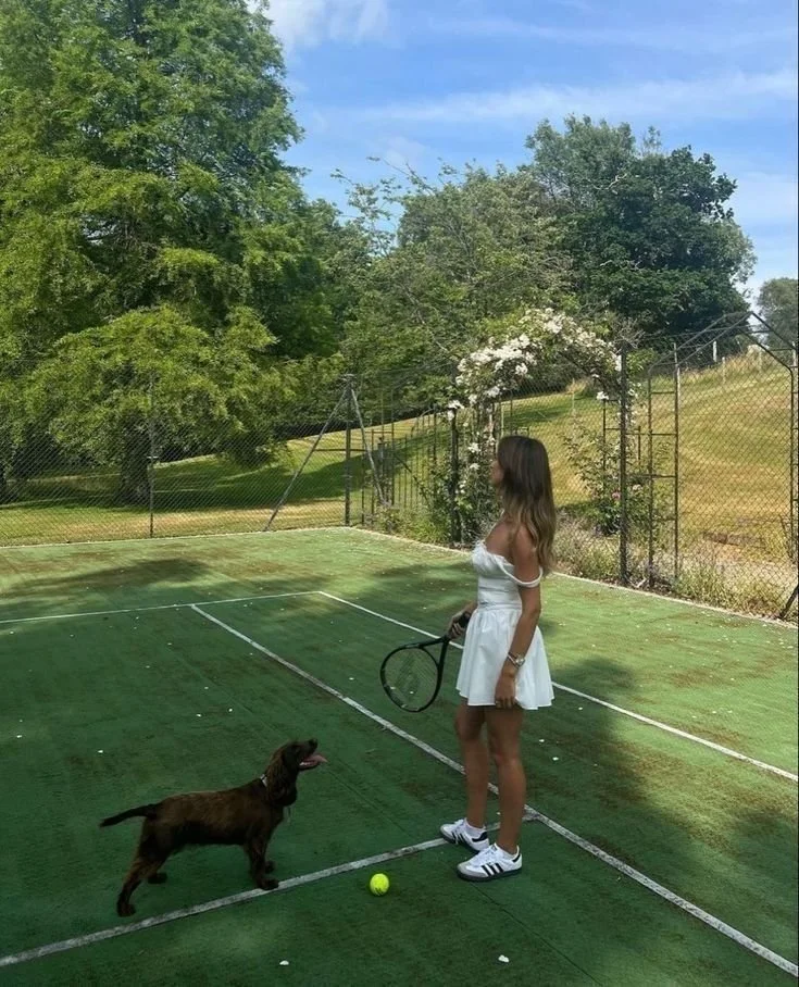 A woman in a white dress and sneakers holding a tennis racket on a grass tennis court, with a brown dog standing nearby. A tennis ball is on the ground, and there are trees and a fence in the background.