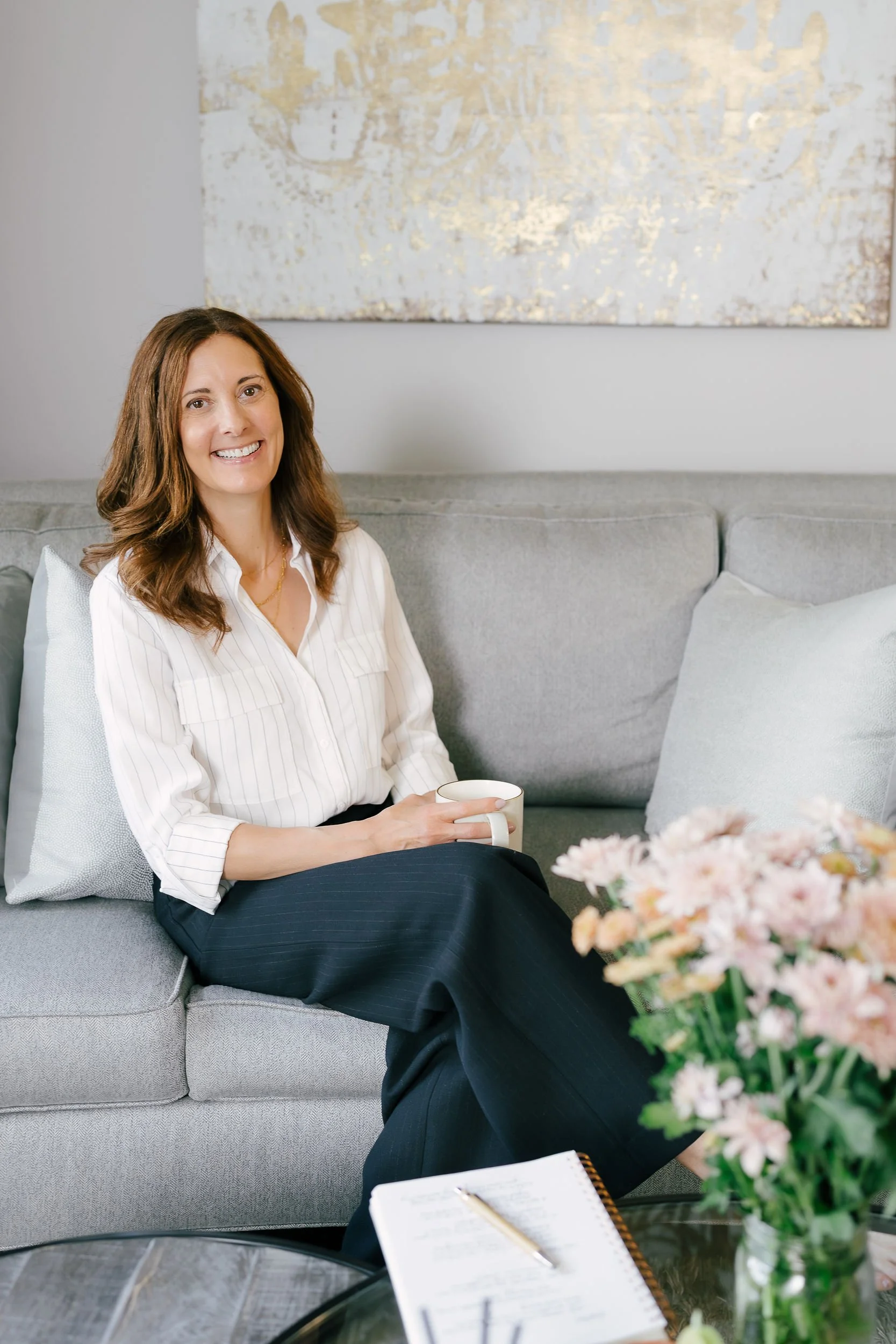 A woman with brown hair is sitting on a gray sofa, smiling, holding a white mug, with a floral arrangement and an open notebook with a pen on a table in front of her.