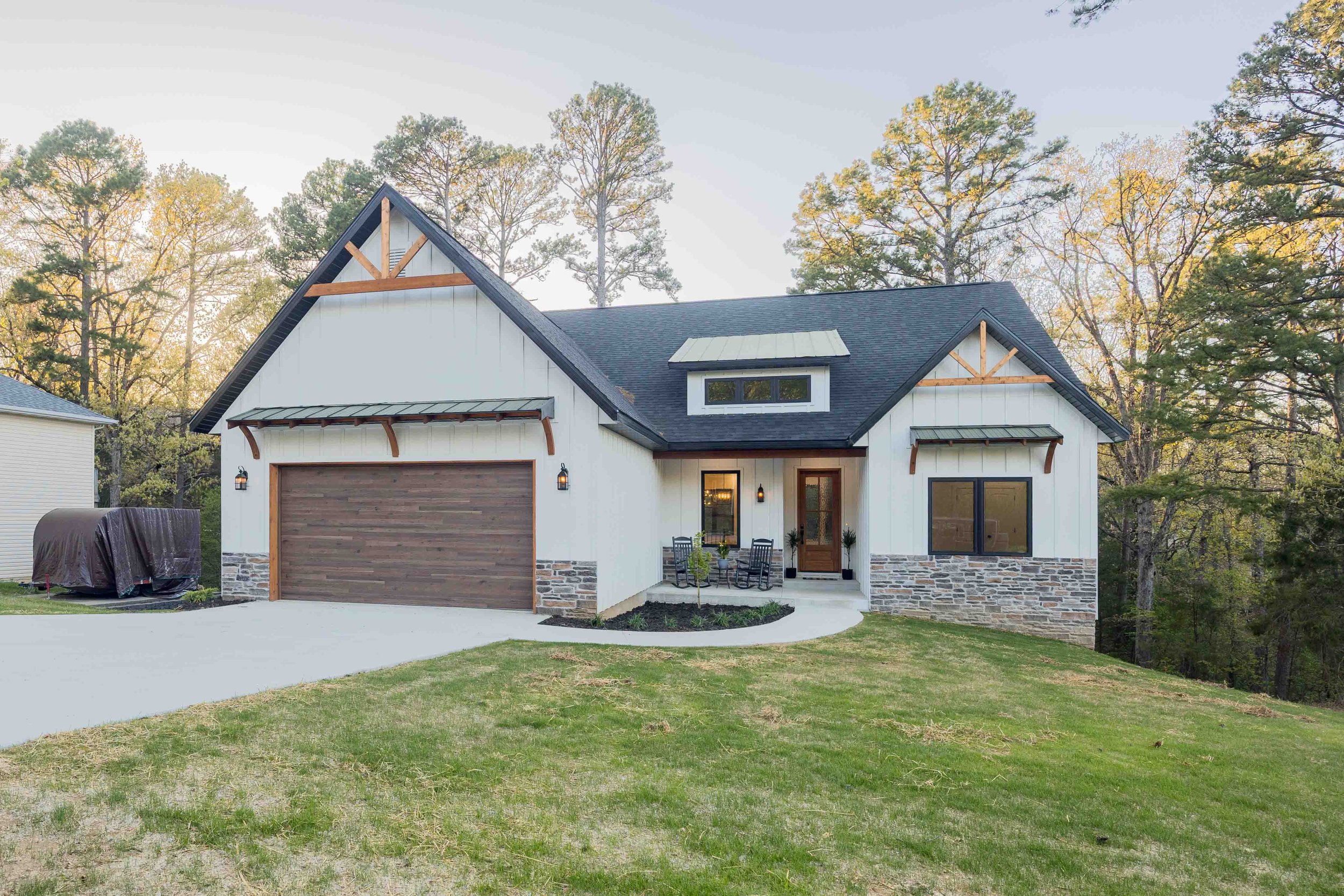 Front view of a modern two-story house with white exterior, black roof, and stone accents, surrounded by tall trees and a lawn.