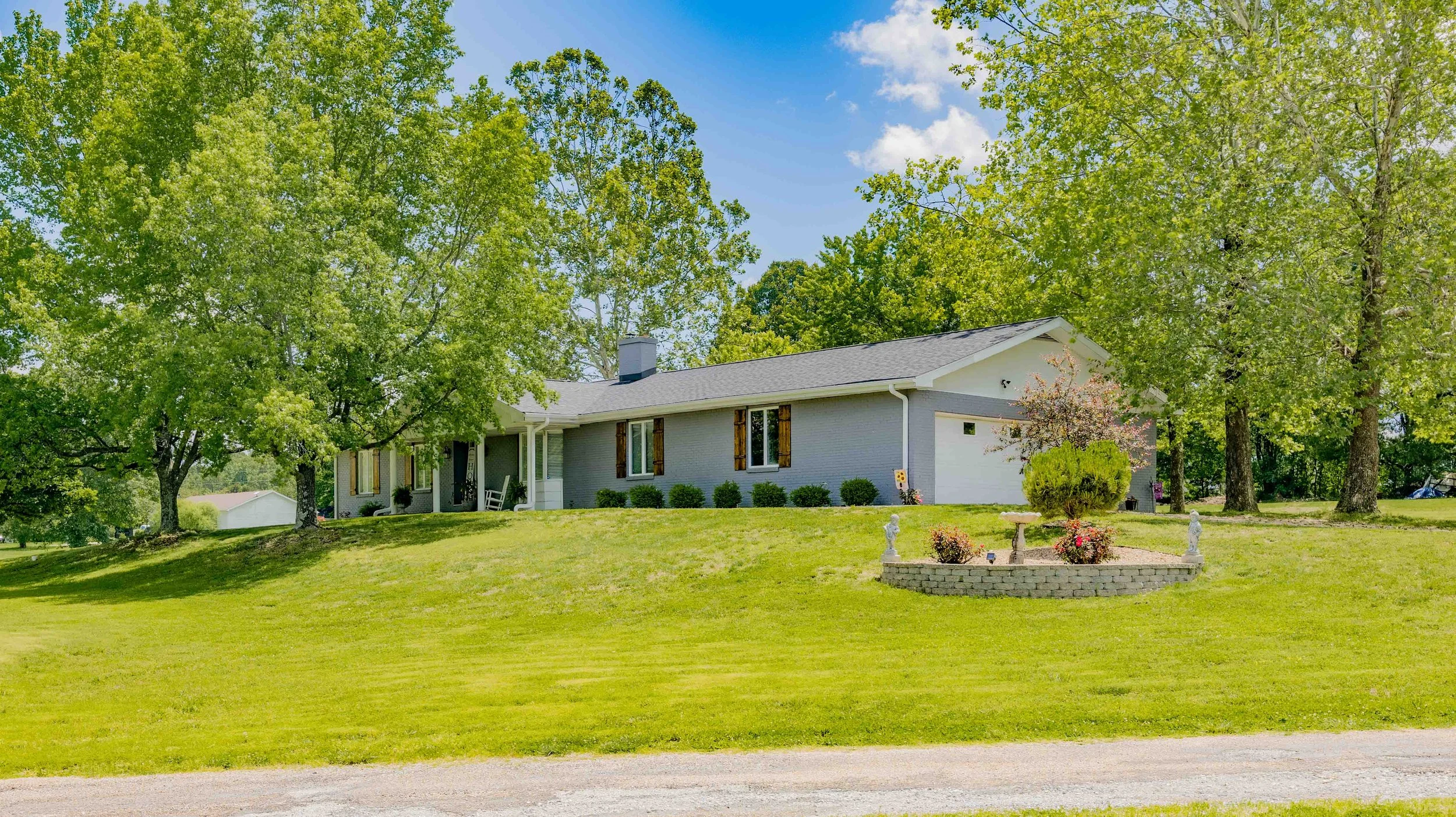 A single-story house with light gray exterior walls and a dark gray roof, surrounded by green trees and a well-maintained front yard with garden ornaments and a circular flower bed.
