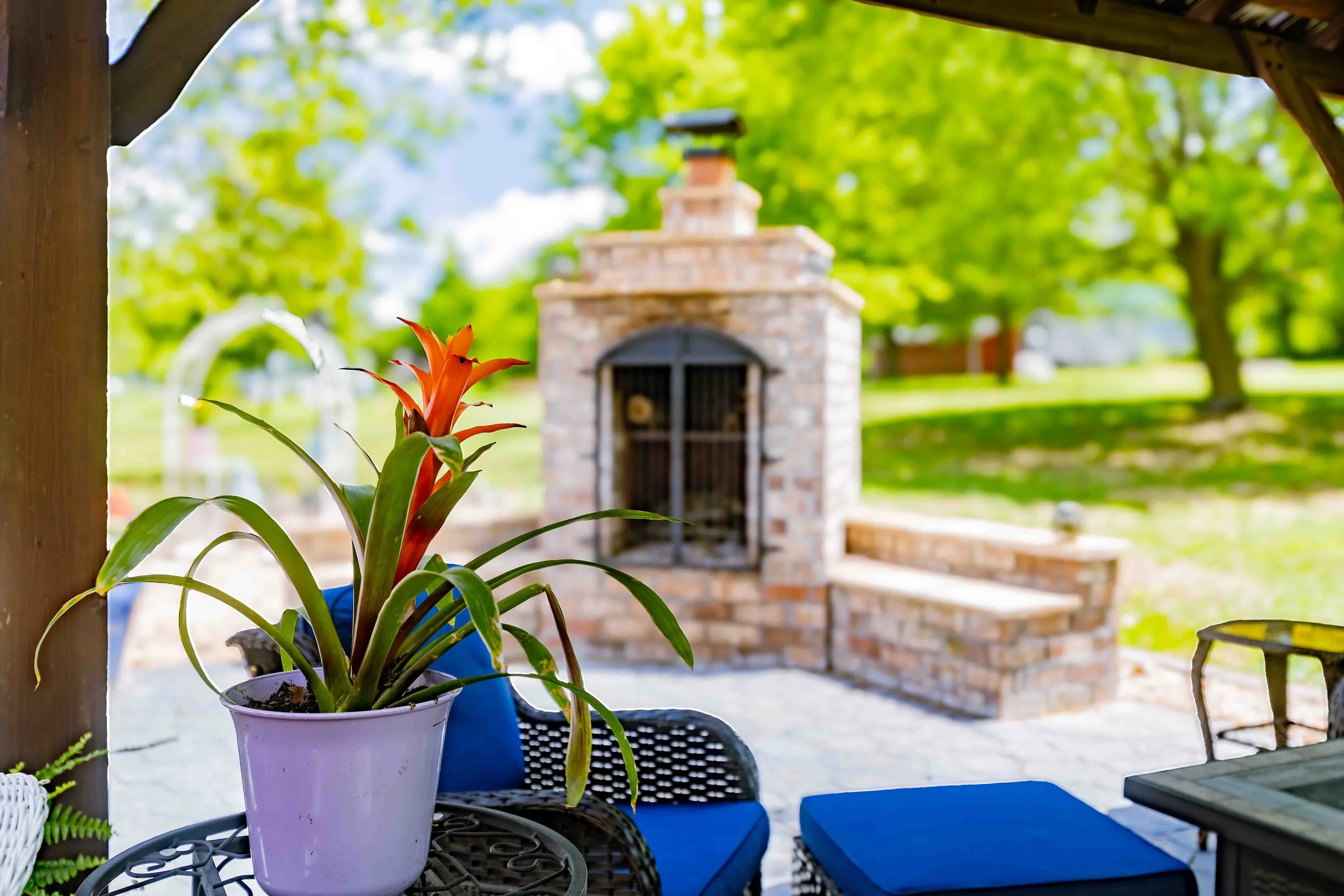 A potted orange flower on a metal table on a porch with an outdoor brick fireplace and trees in the background.