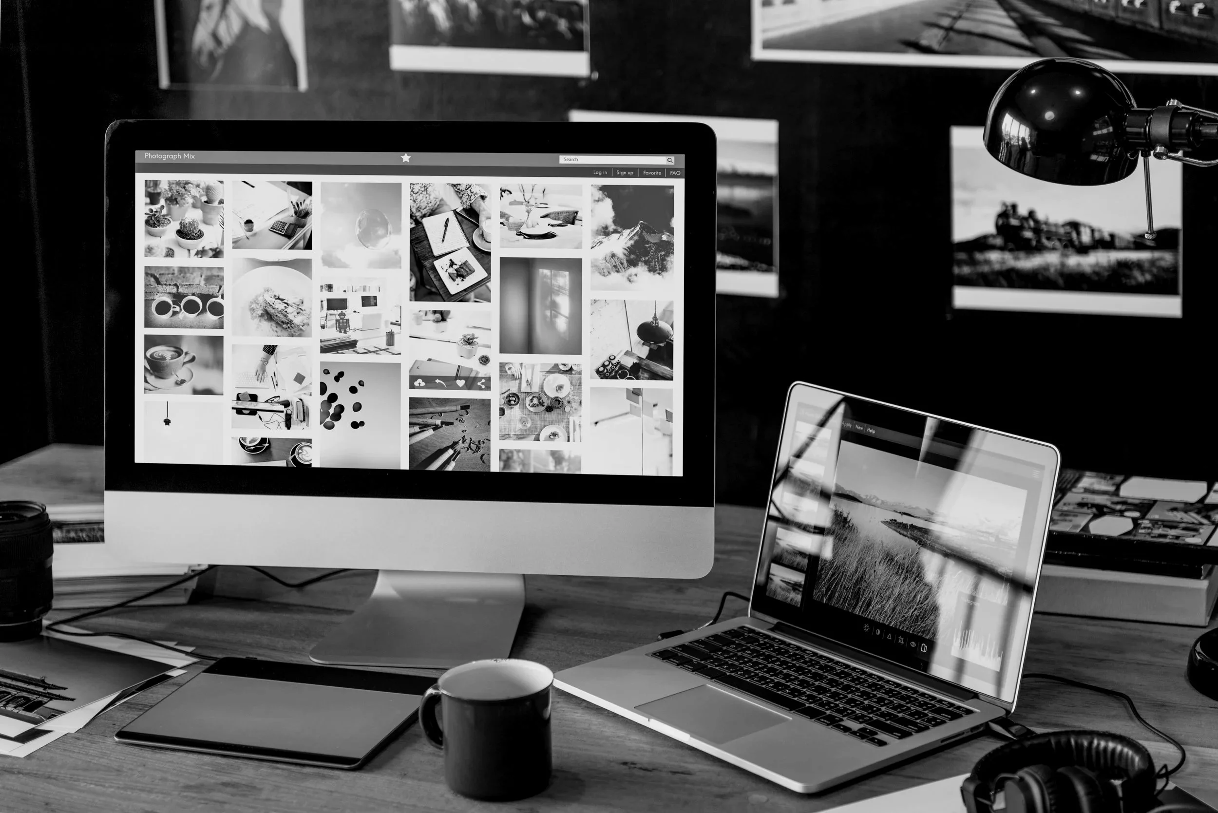 A workspace with a large desktop monitor displaying a grid of black and white photographs, a laptop showing a landscape image, a coffee mug, headphones, and various papers scattered on a wooden desk.