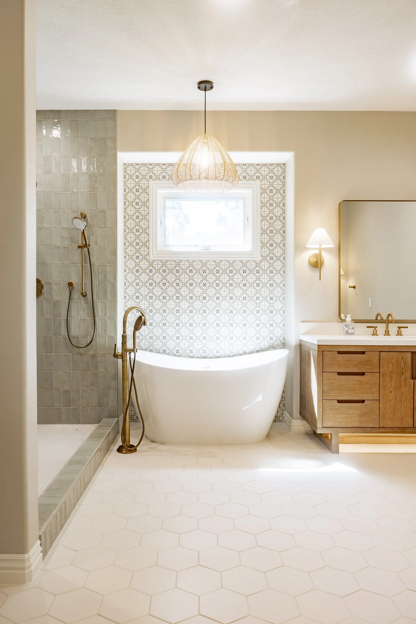 Modern bathroom featuring a walk-in shower with beige tiles and a brass showerhead, a freestanding bathtub against patterned wall tiles under a small window, and a wooden vanity with a round mirror and wall-mounted light fixture.
