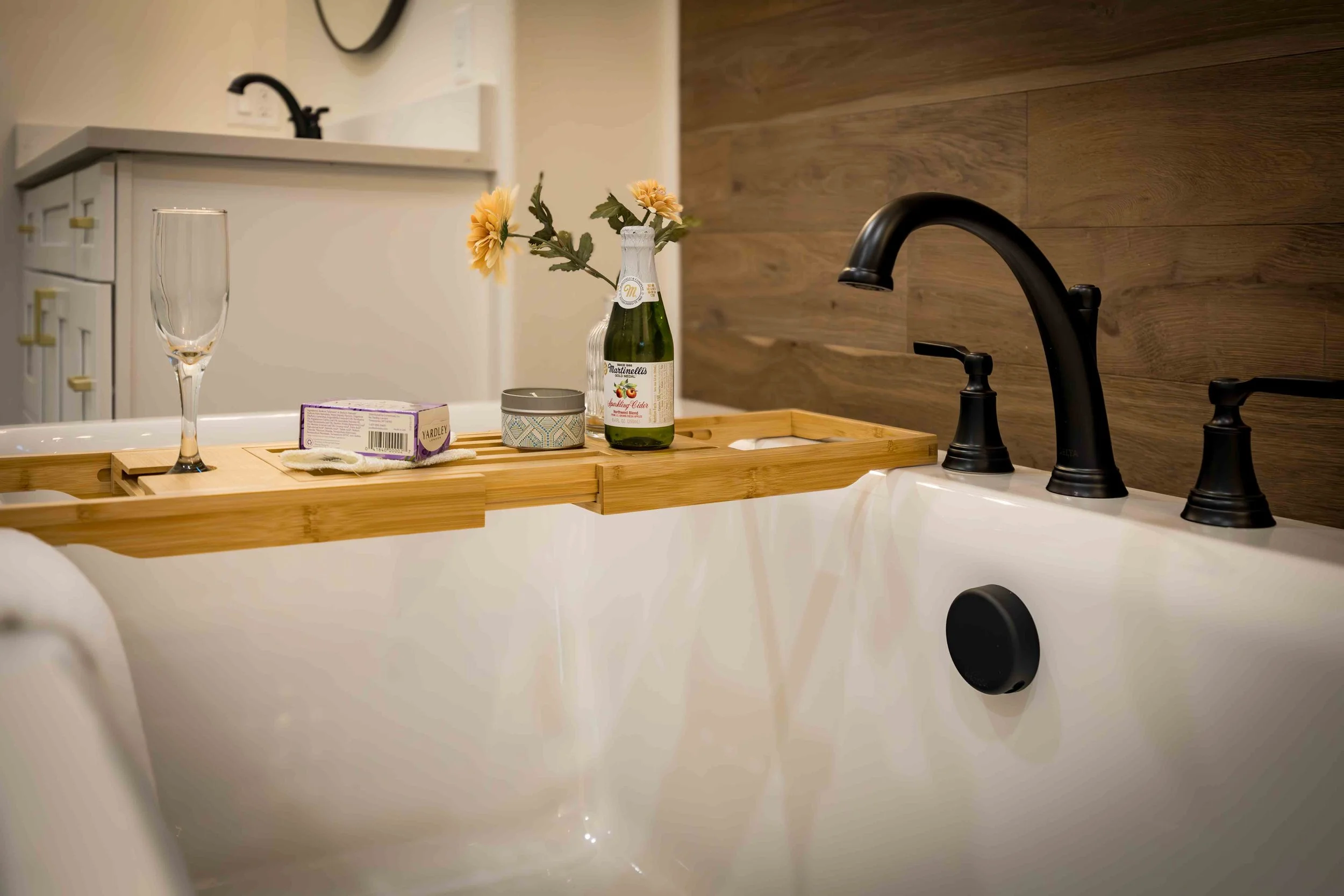 A bathroom with a bathtub, a black faucet, a wooden bath tray holding a wine glass, a box, a small jar, a vase with yellow flowers, and a bottle of sparkling cider, with a wooden accent wall in the background.