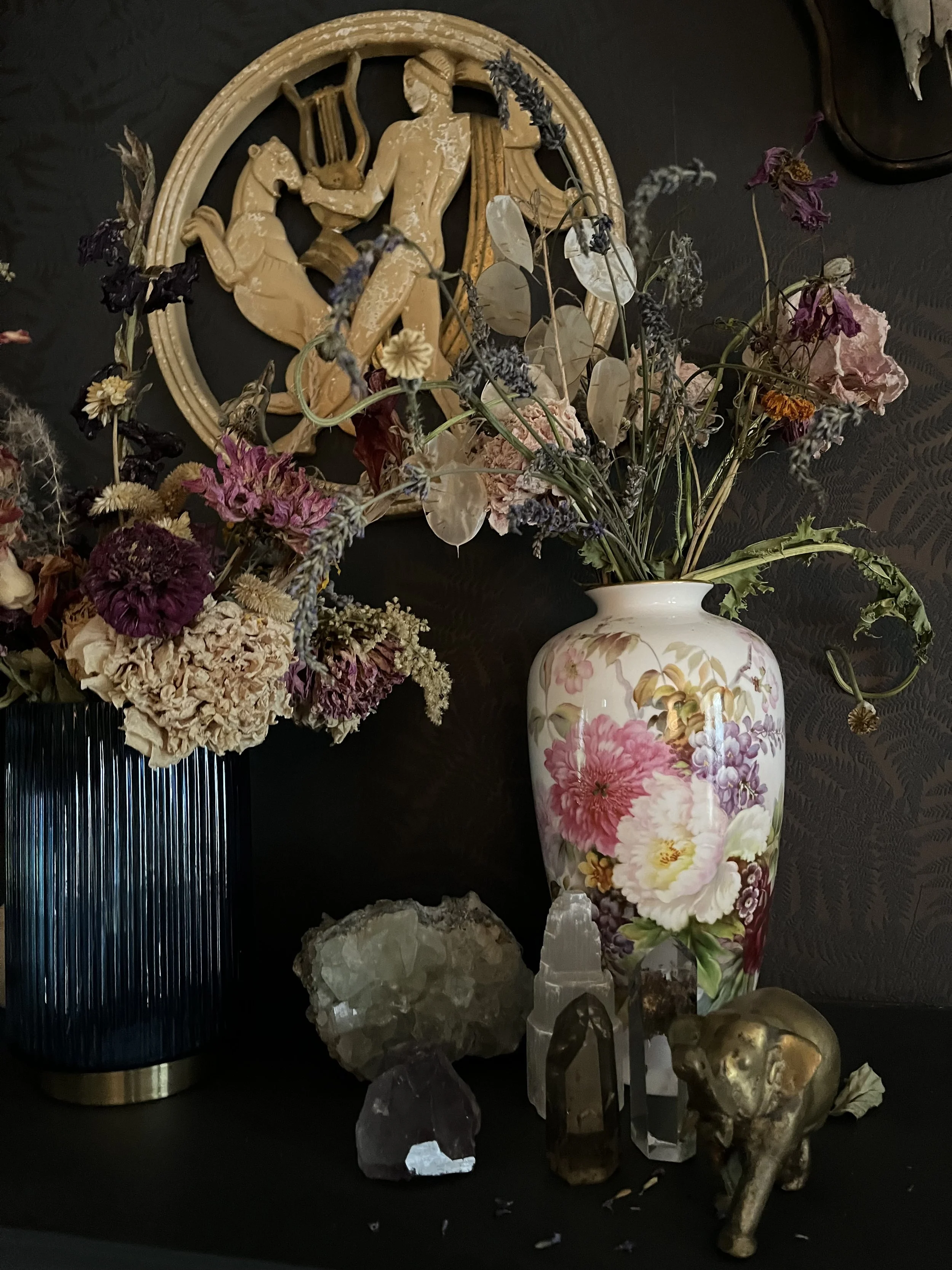 Decorative arrangement with dried flowers in vases, a relief carving of a figure with a lyre, crystals, and a small elephant figurine.