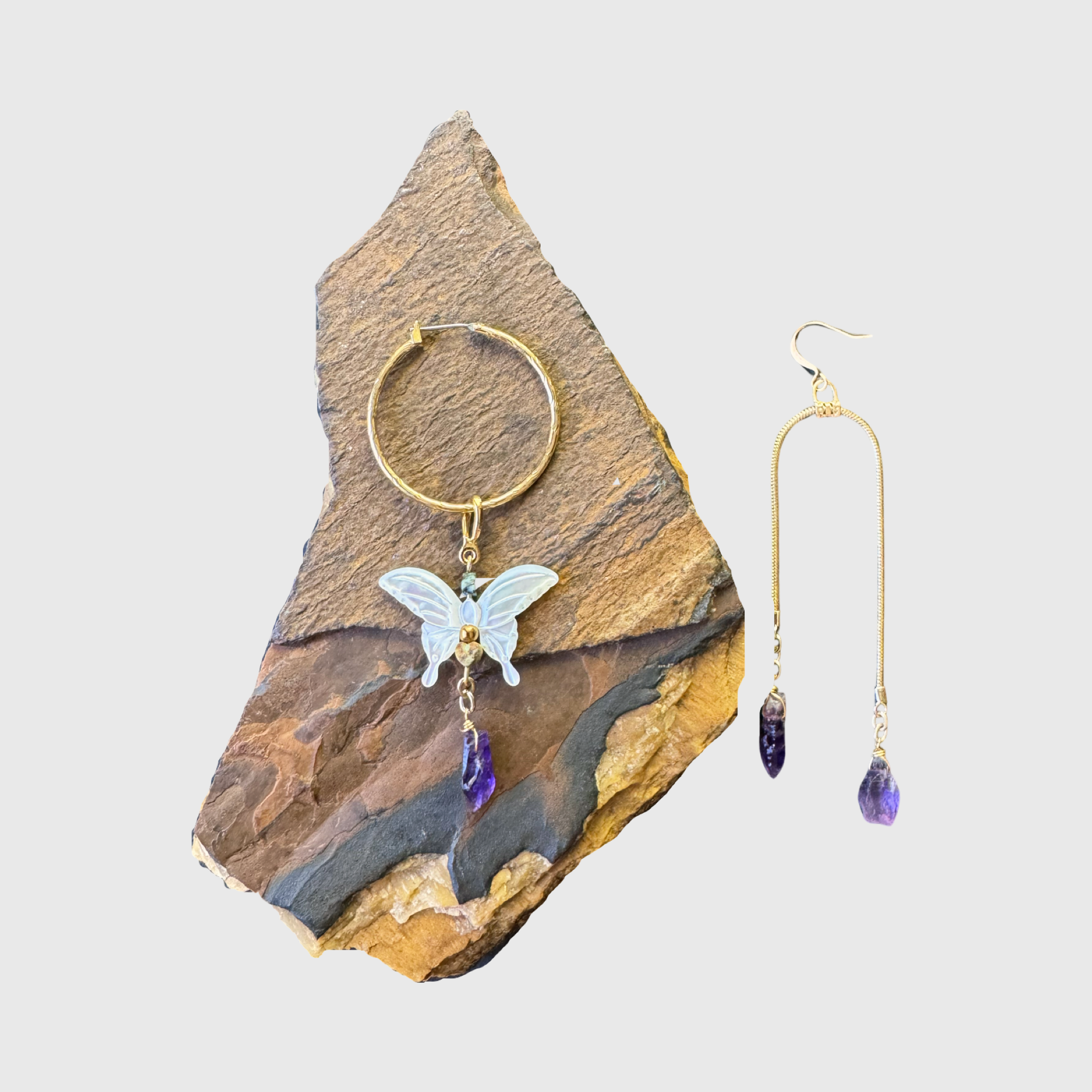 Butterfly charm hoop earring and long chain earring with amethyst stones on a rock background.