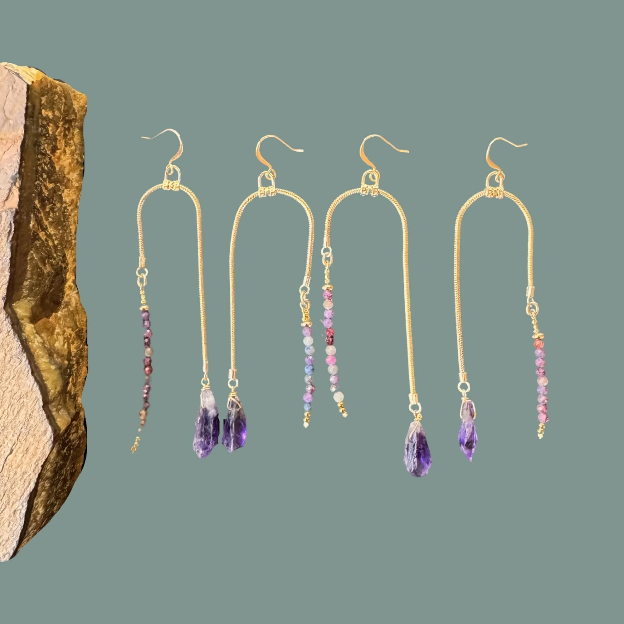Four pairs of dangling gold wire earrings with colorful bead detailing and amethyst crystal points, displayed on a green background next to a piece of rough rock.