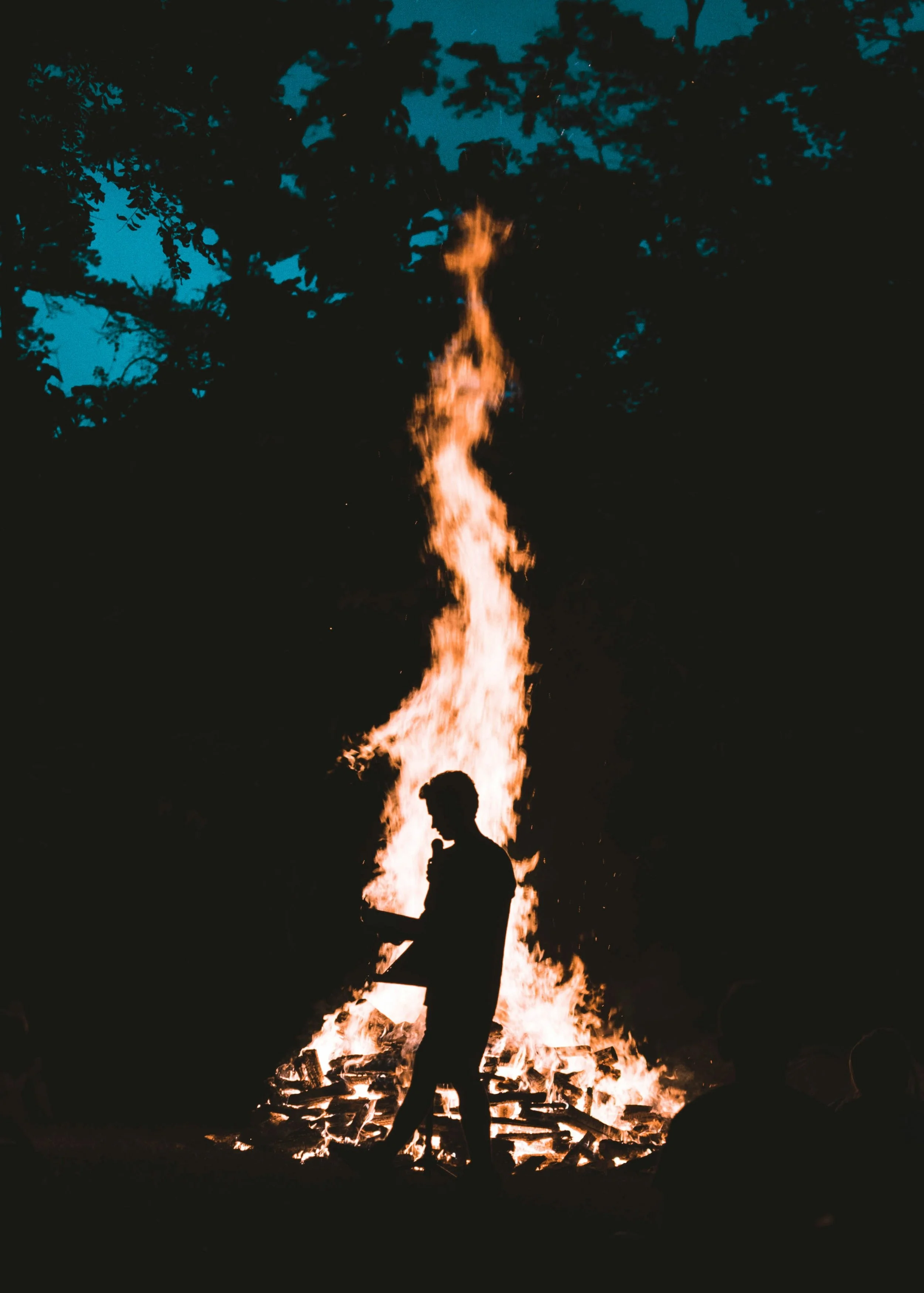 Silhouette of a person standing near a large outdoor fire during nighttime, with trees visible in the background.