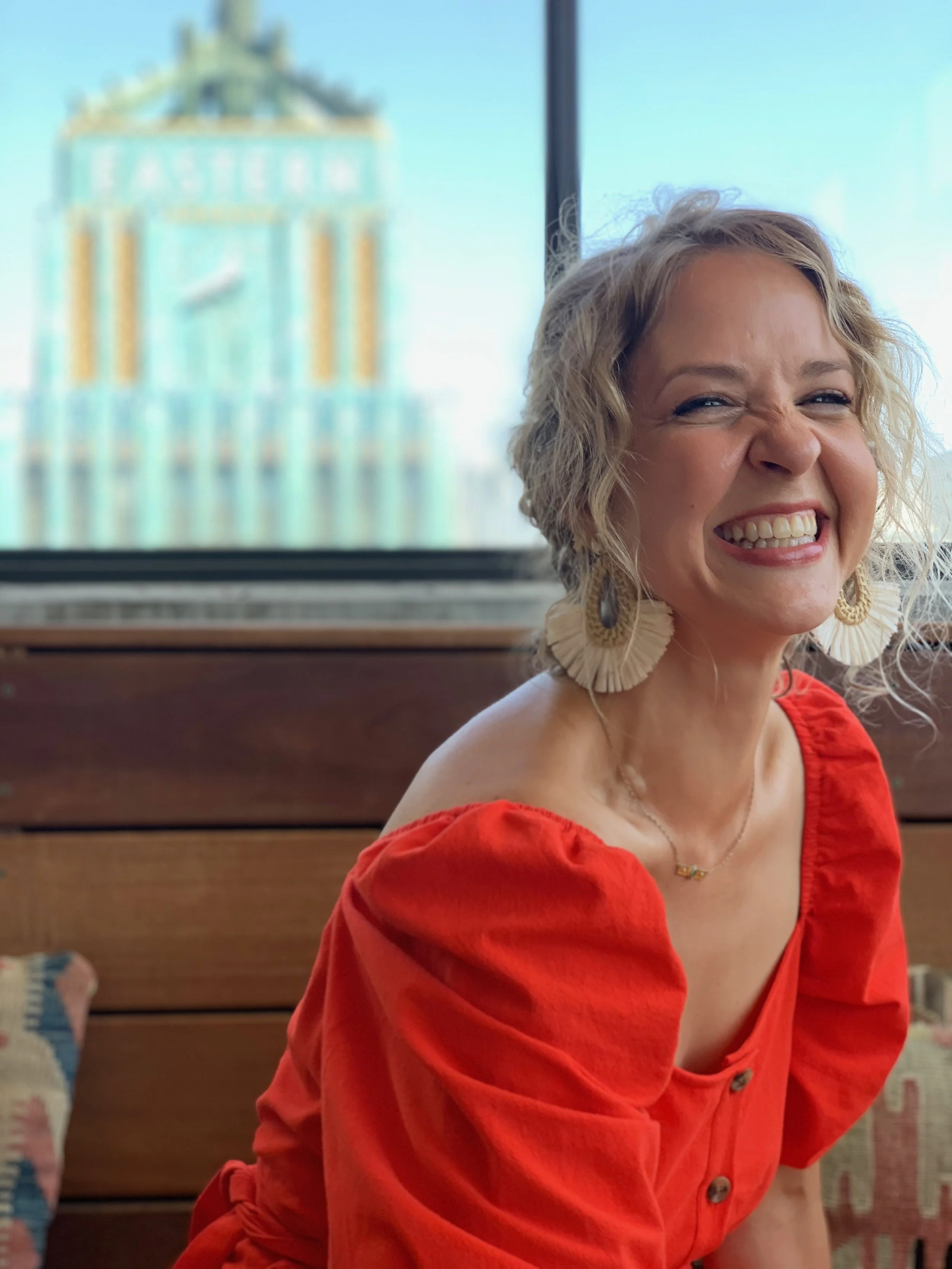 A smiling woman with curly blonde hair, gold earrings, red dress with puffed sleeves, and a necklace, sitting in front of a window with a blurred cityscape in the background.
