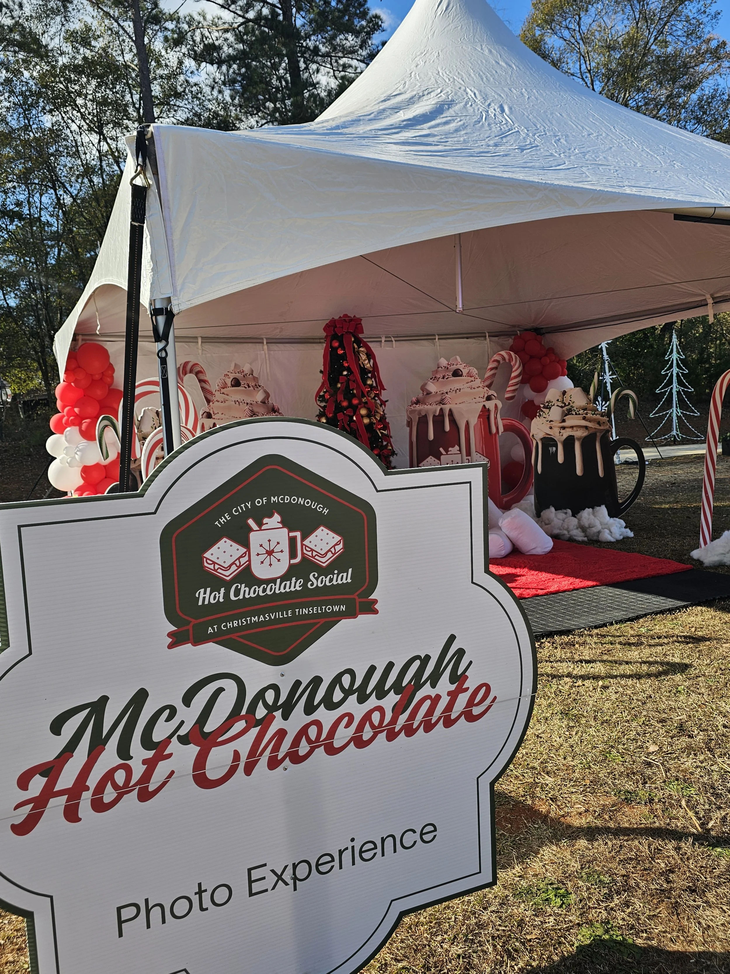A festive outdoor photo setup for McDonough Hot Chocolate, featuring large decorated mugs of hot chocolate, candy canes, cotton snow, and Christmas decorations inside a white tent with a sign in front promoting the photo experience.