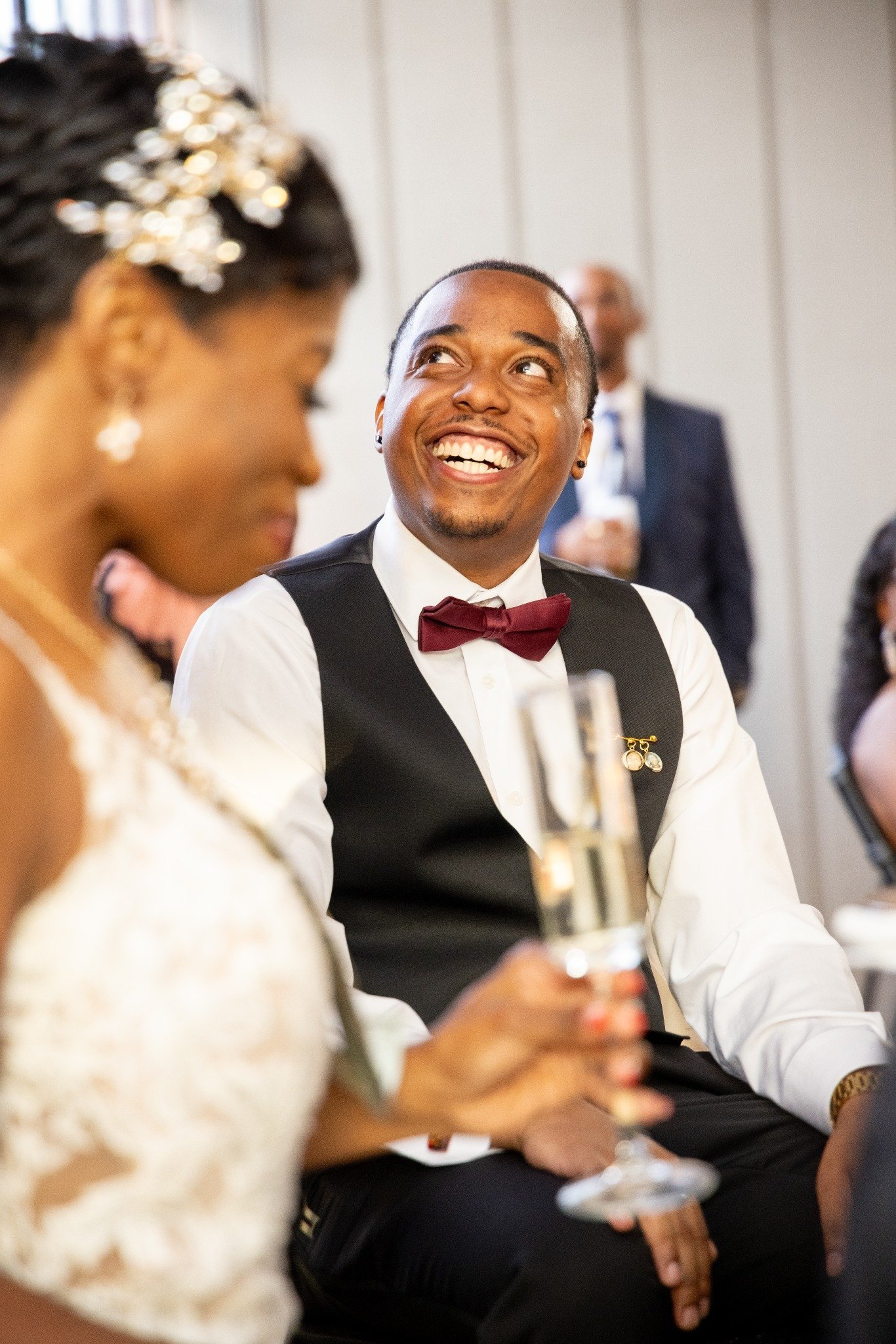 A man in a tuxedo with a bow tie smiling at a woman in a wedding dress during a celebration, holding a glass of champagne.