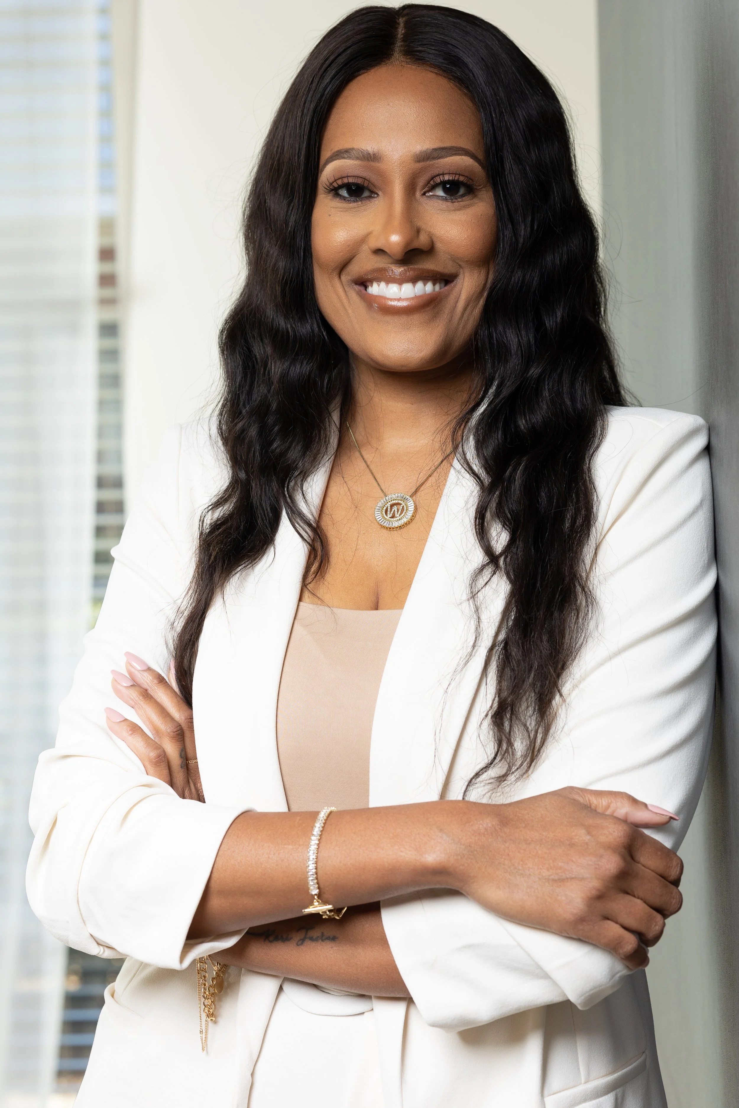 A confident woman with long, wavy black hair wearing a white blazer, beige top, and jewelry, standing with arms crossed and smiling indoors.