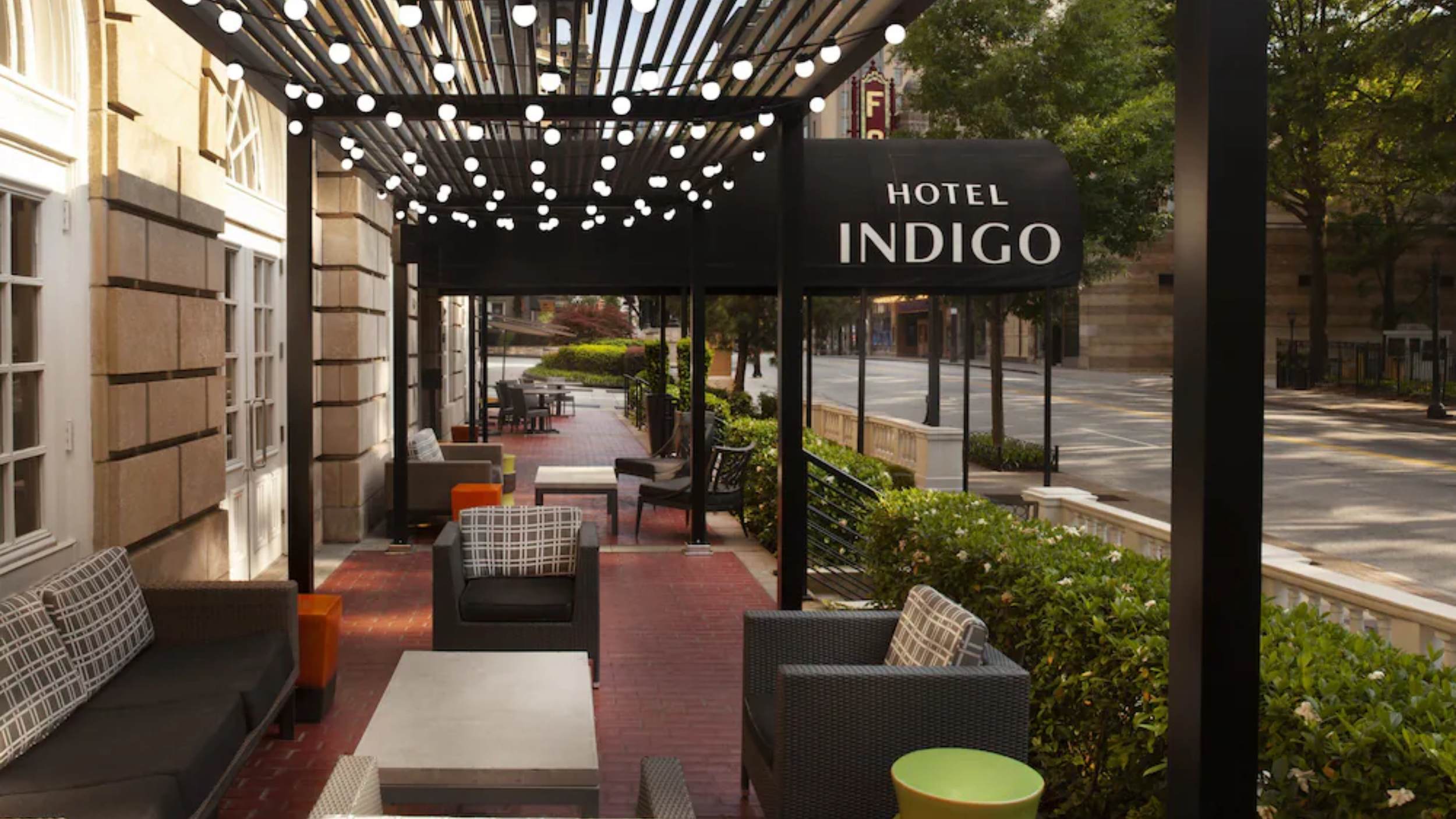 Outdoor seating area of Hotel Indigo with chairs, tables, and string lights under a black awning with the hotel name.