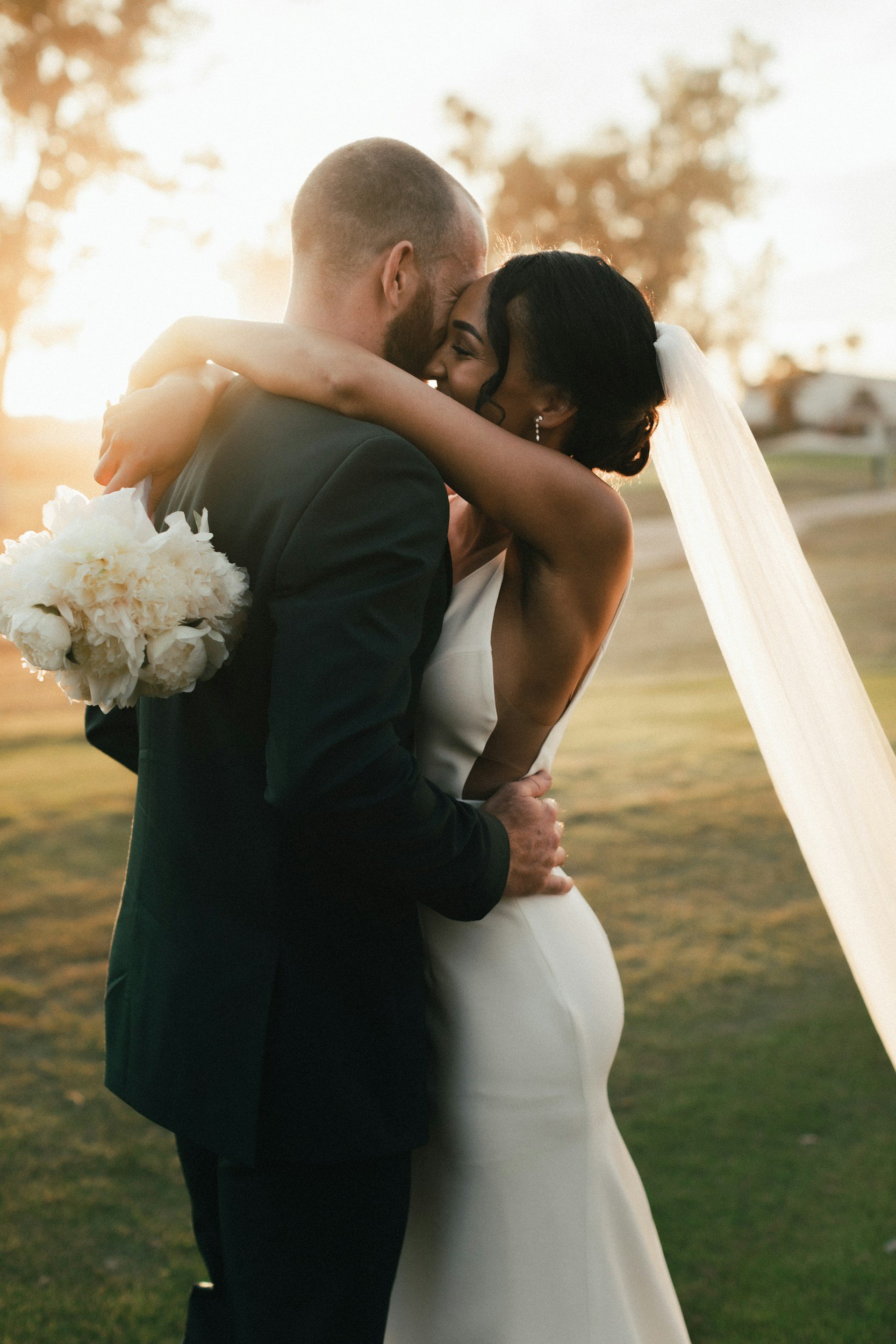 A bride and groom hugging during their outdoor wedding at sunset, with the bride holding a bouquet of white flowers.