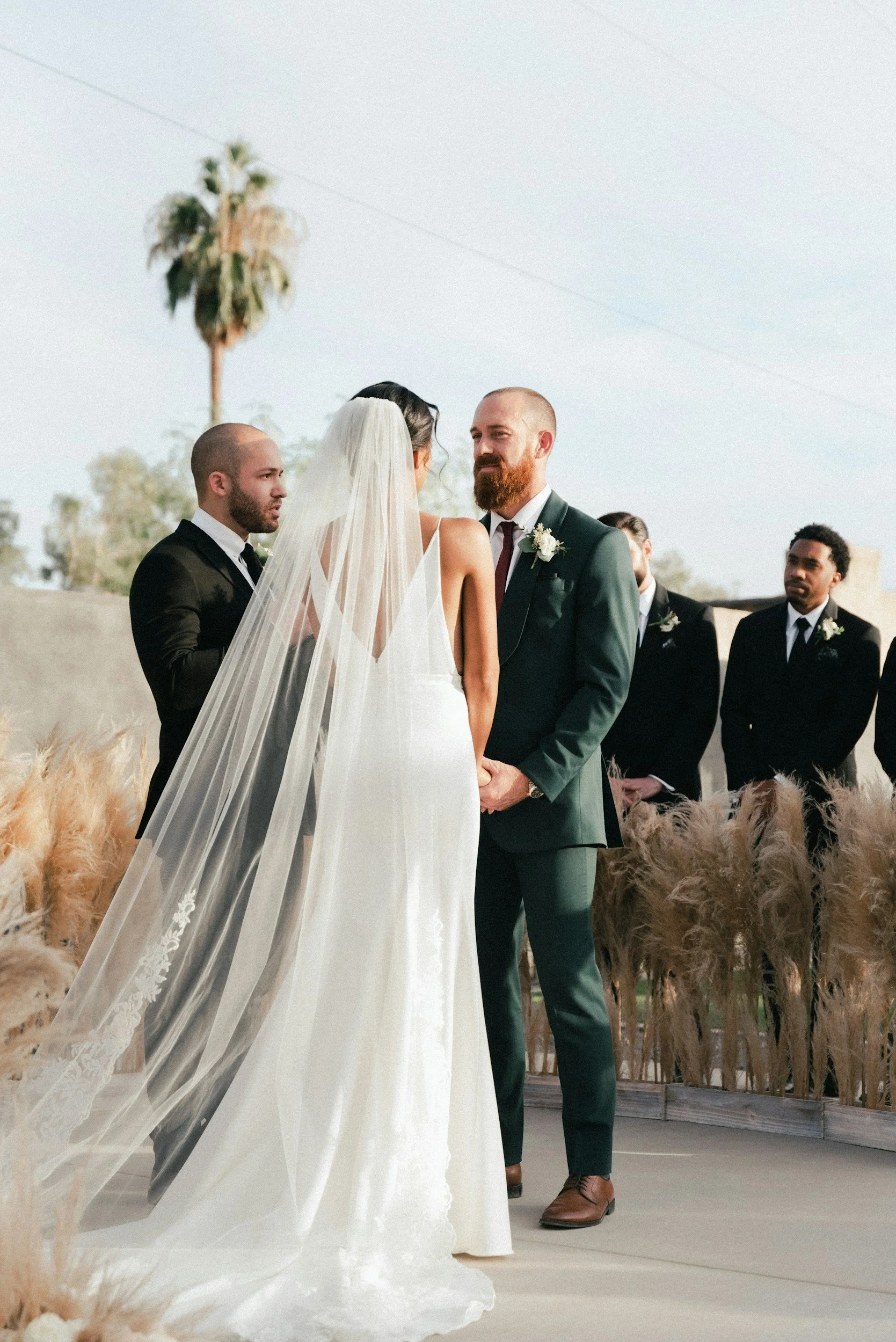 Bride and groom exchanging vows outdoors, with bridesmaids and groomsmen in the background, and a palm tree in the distance.