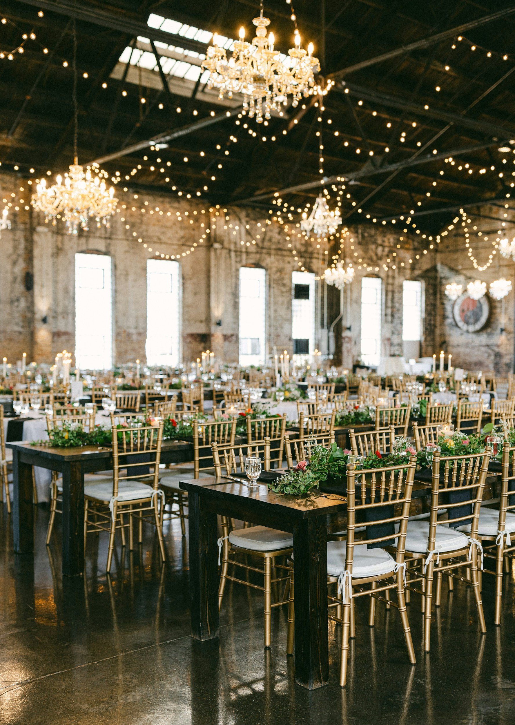 Elegant event space with exposed brick walls, large windows, chandeliers, and string lights. Long tables with dark wood surfaces are decorated with green foliage and floral centerpieces surrounded by gold chairs with white cushions.