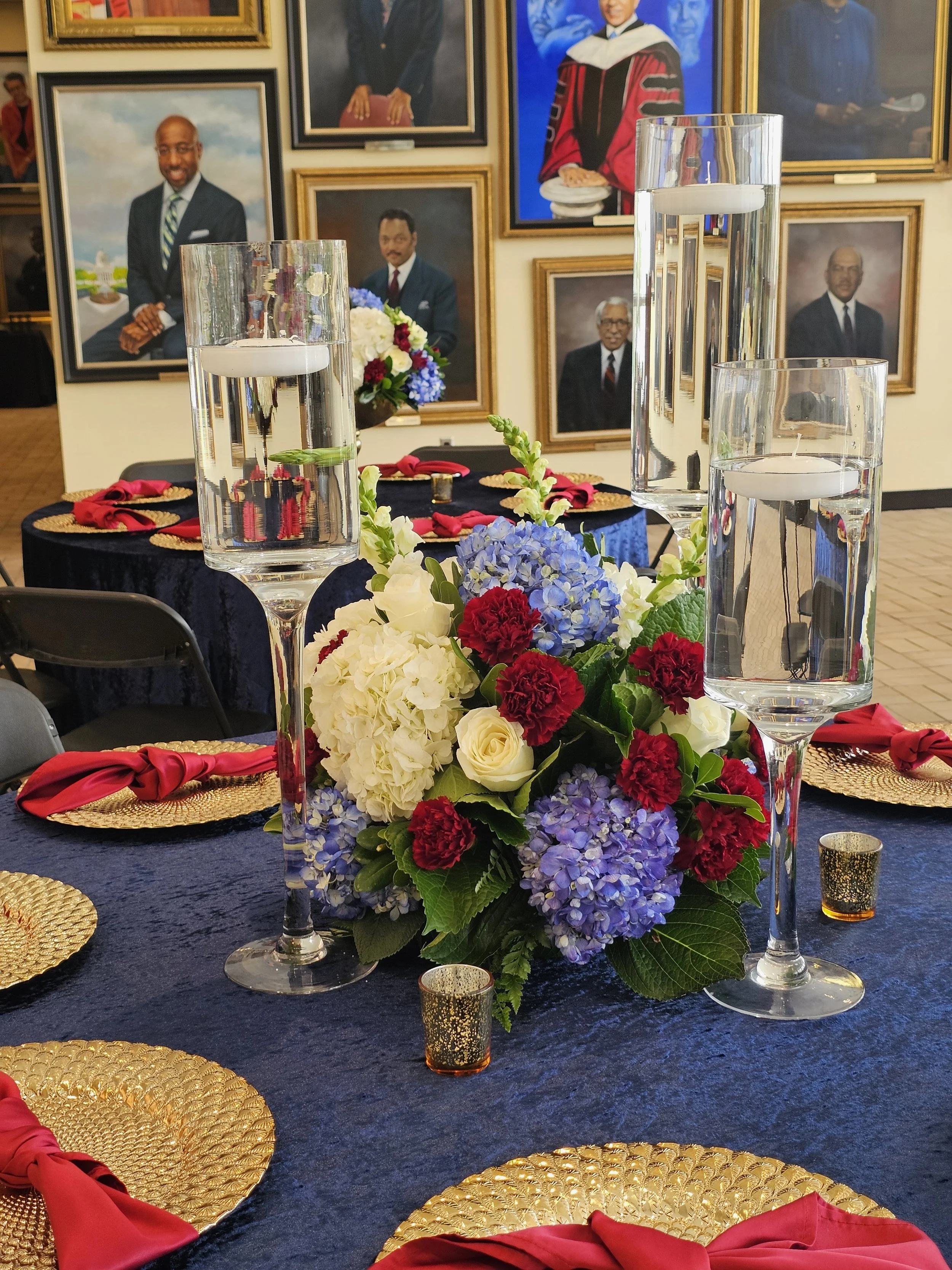 Elegant dining table setup with a floral centerpiece of white, red, and purple flowers, tall glass candle holders with floating candles, gold placemats with red napkins, and a background of framed portraits of African American leaders.