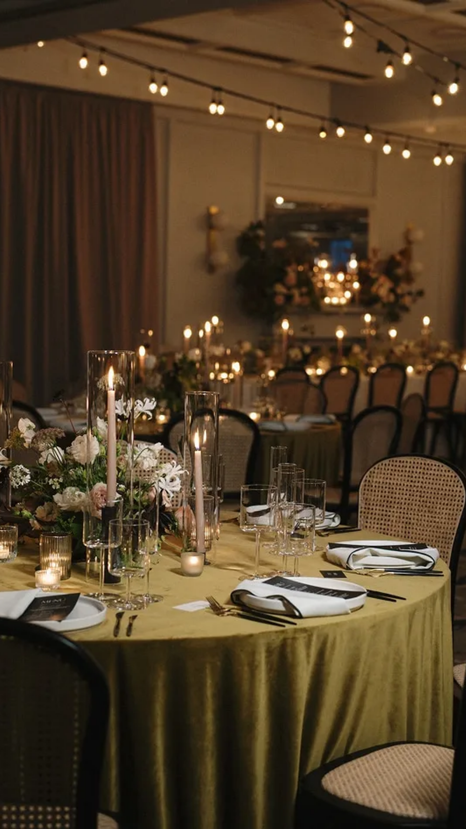 Elegant banquet table decorated with candles and floral arrangements in a dimly lit event hall with string lights overhead.