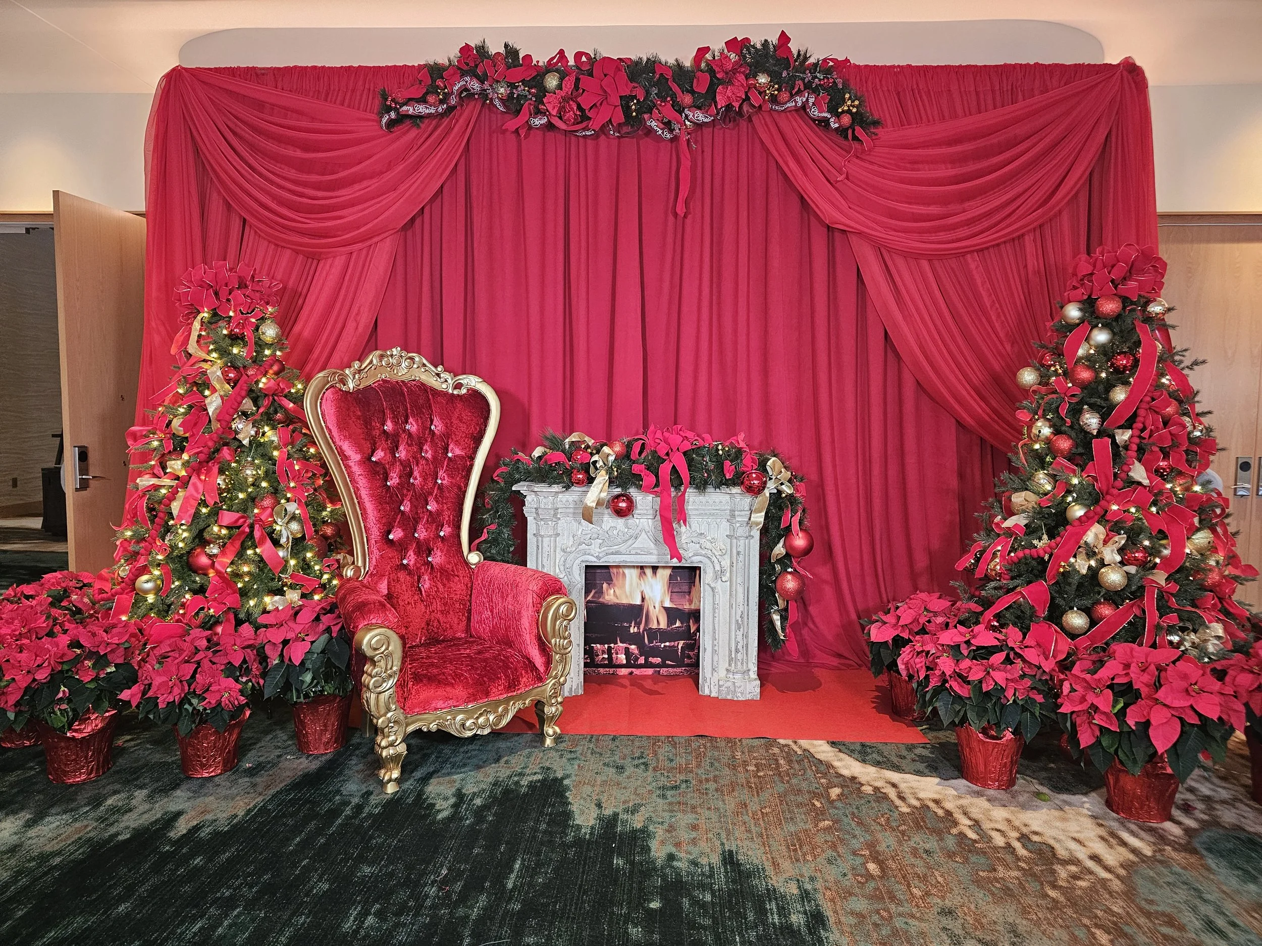 Festive Christmas decorations with a red velvet throne, decorated Christmas trees, poinsettias, a fireplace, and a pink draped backdrop.