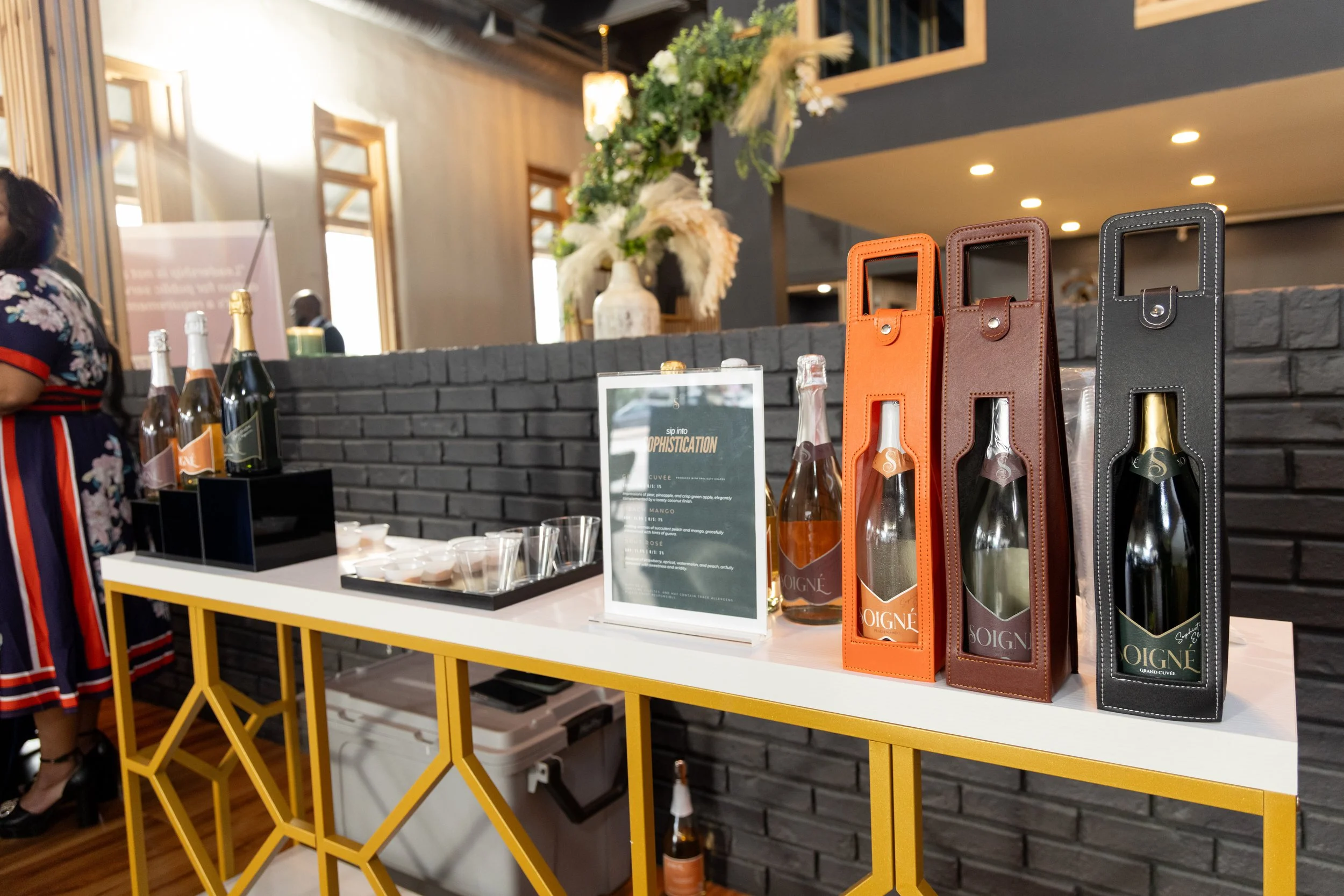 Display of bottles of champagne or sparkling wine in colorful holders on a white and gold table at an event or tasting, with decorative flowers and people in the background.