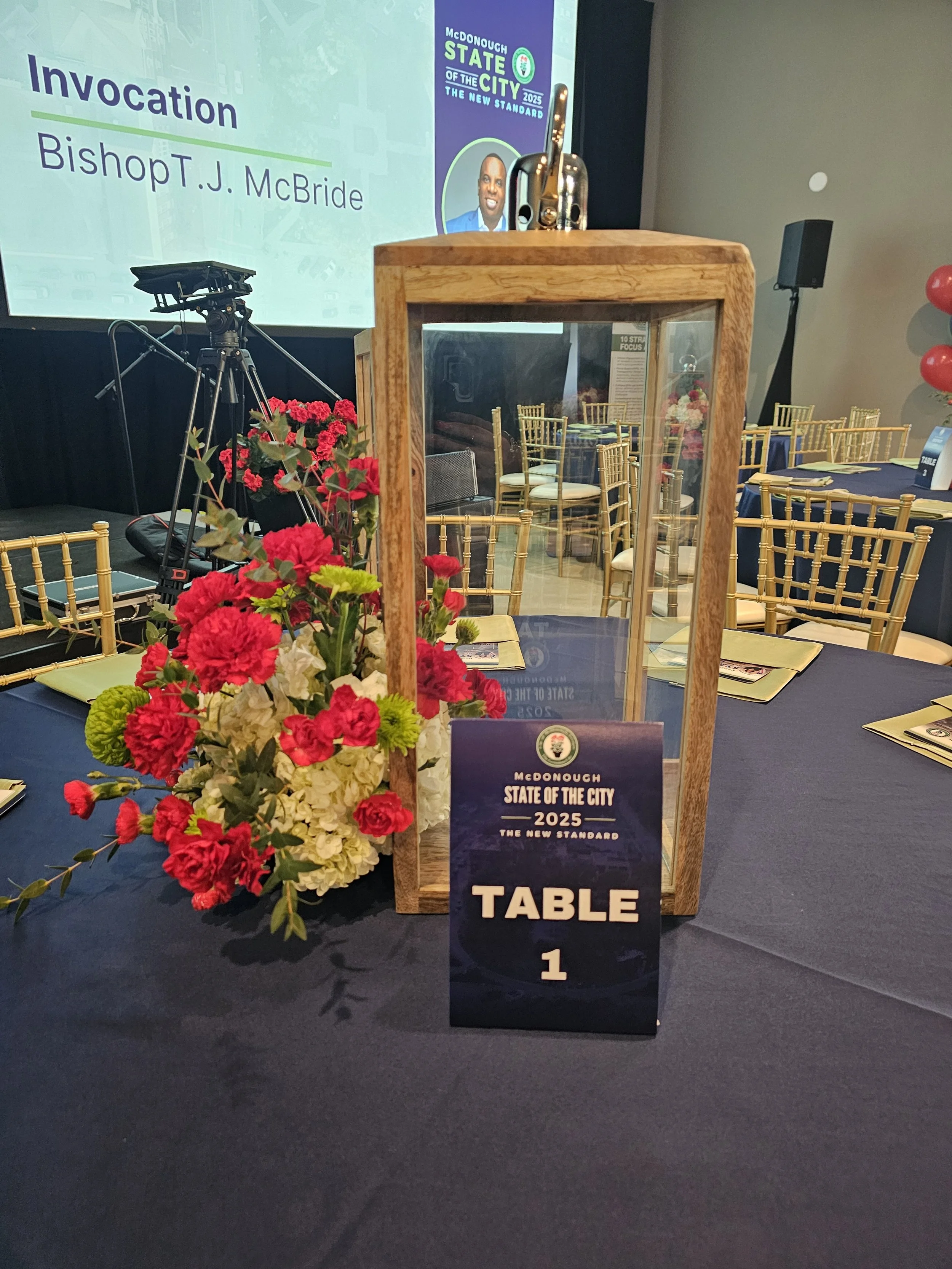 A table at an event with a floral arrangement, a wooden and glass display case, and a sign reading 'Table 1' in front of a screen showing information about Bishop J. McBride and the 2025 McDonough State of the City event.