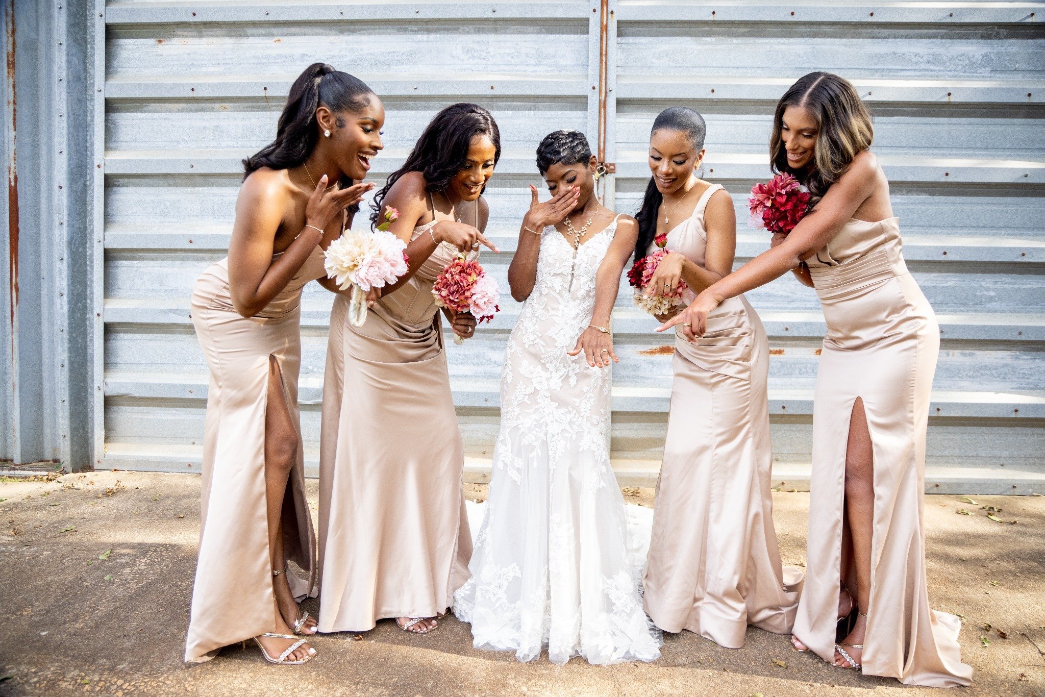 A bride in a white lace wedding dress showing her engagement ring, surrounded by her five bridesmaids in matching champagne-colored dresses with high slits, all holding pink and white bouquets, excitedly looking at the bride's ring.