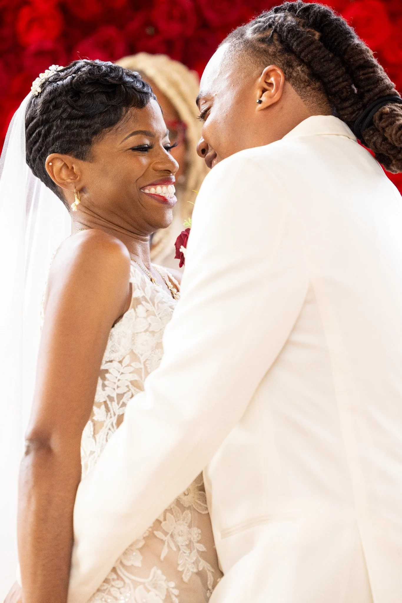 A bride and groom smiling and looking at each other during their wedding ceremony, with red floral decorations in the background.