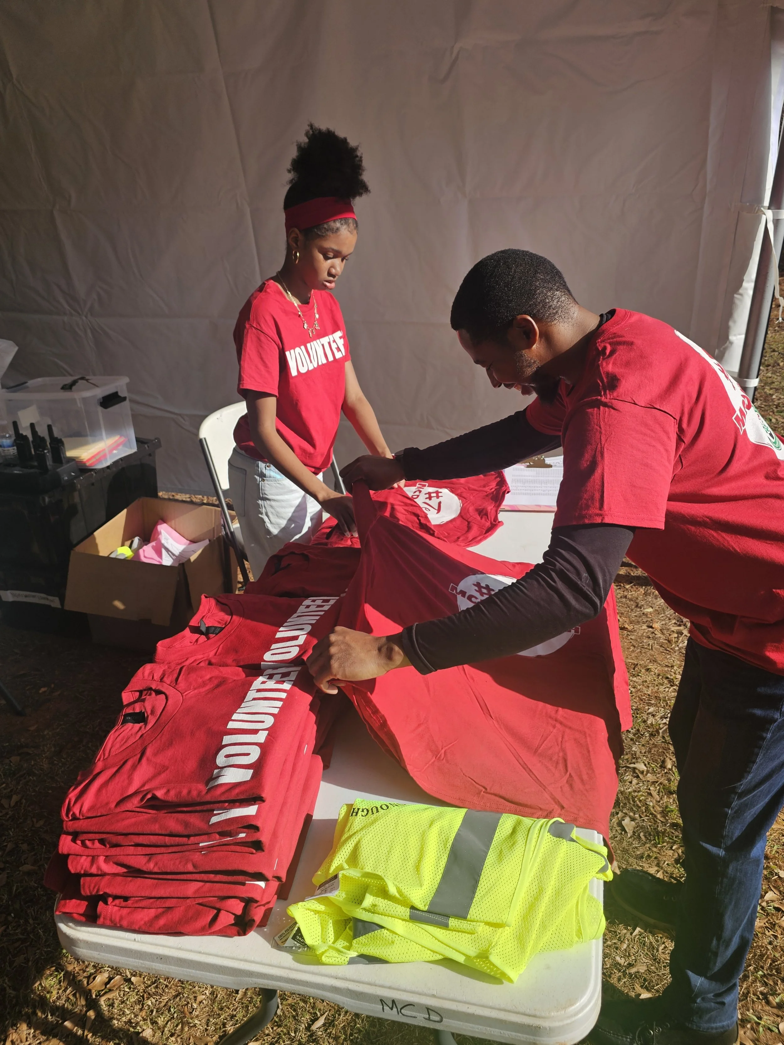 Two volunteers organizing red t-shirts with the word 'VOLUNTEER' printed on them and a yellow reflective safety vest on a folding table outside under a tent.