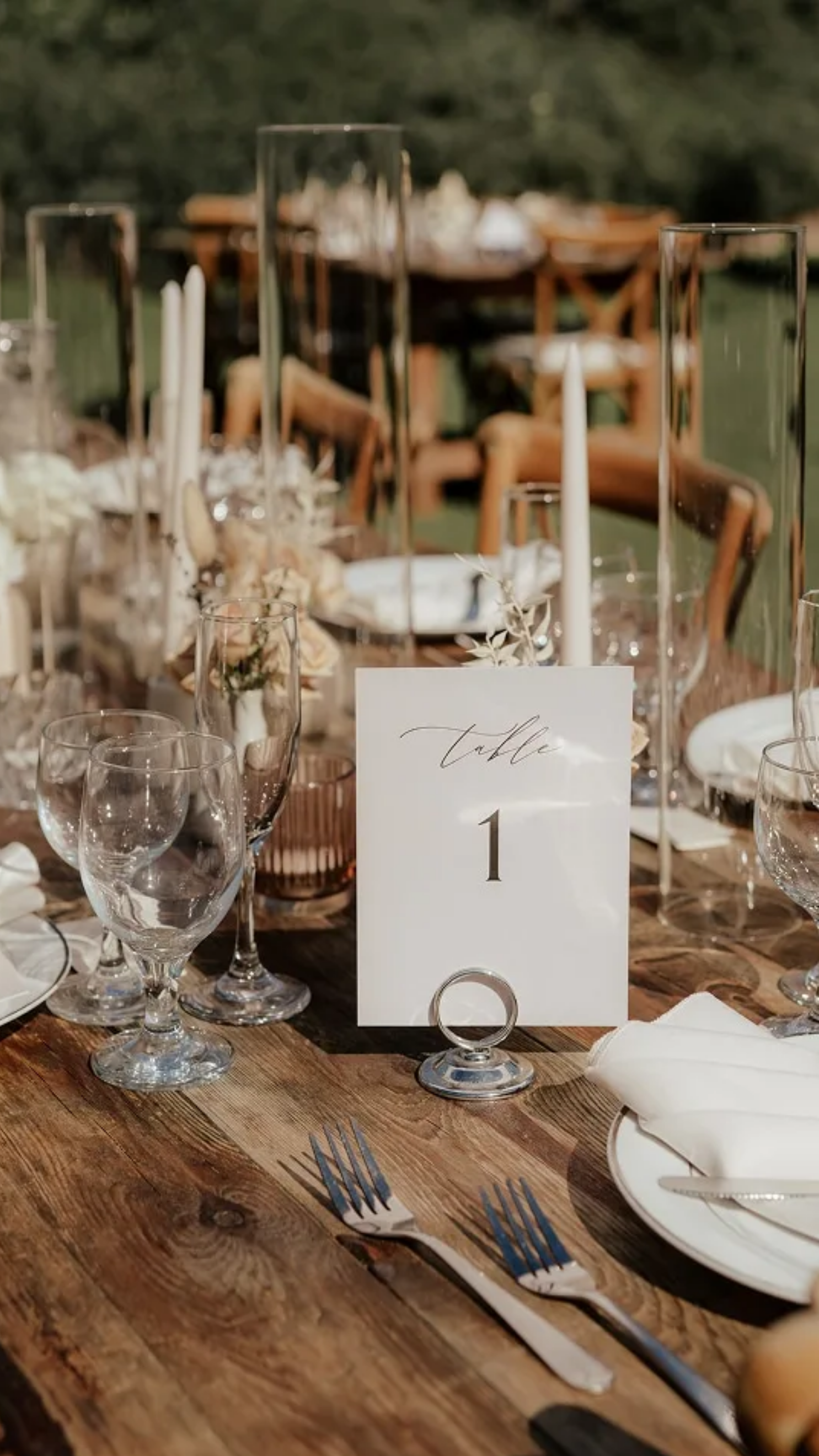 Elegant outdoor wedding reception table set with glassware, candles, flower arrangements, and a table number card displaying 'Table 1' on a rustic wooden table.