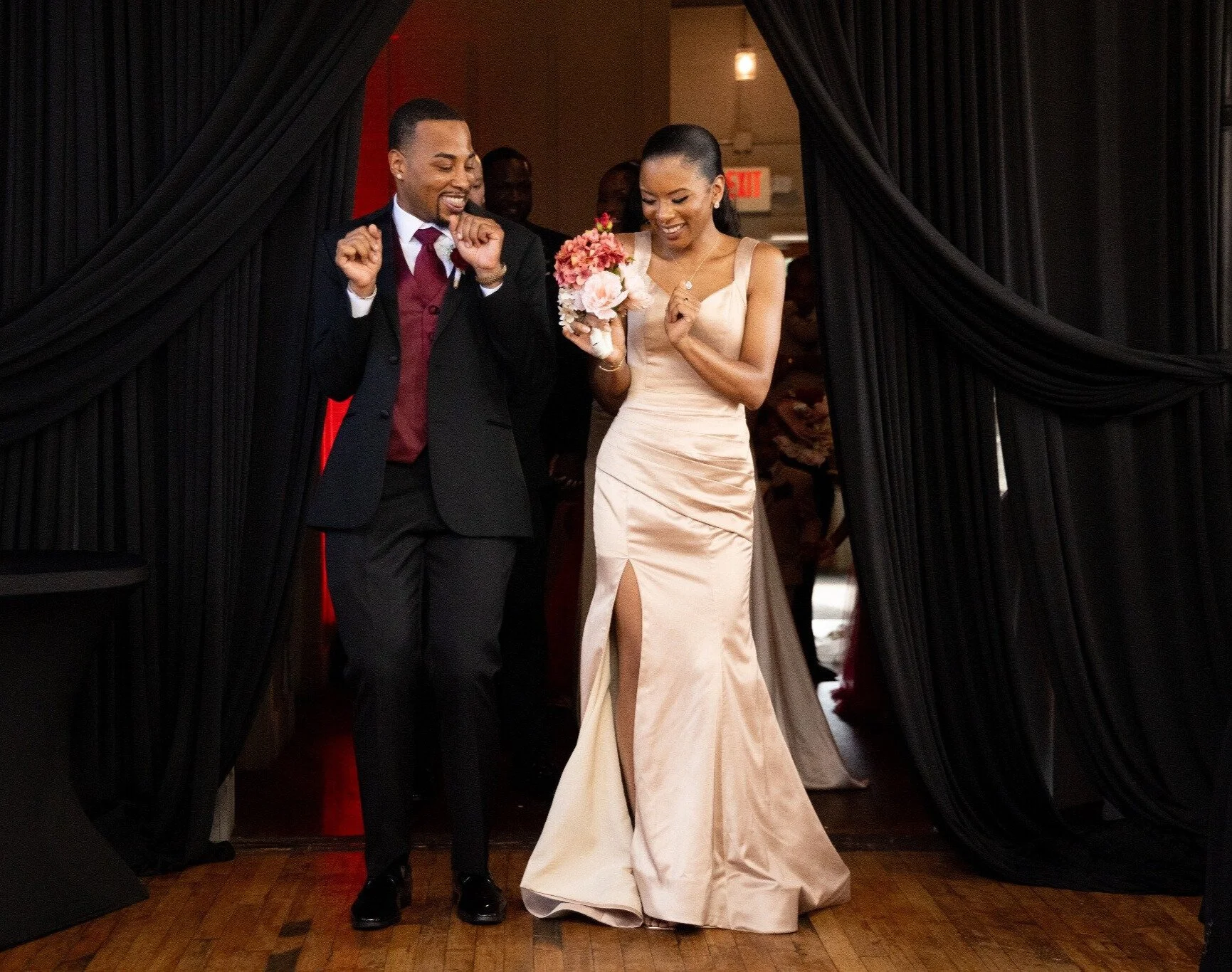 A newlywed couple walking through a black curtain at their wedding reception, smiling and celebrating. The bride is holding a bouquet of pink and white flowers and wearing a satin gown with a slit. The groom is wearing a black suit with a maroon vest
