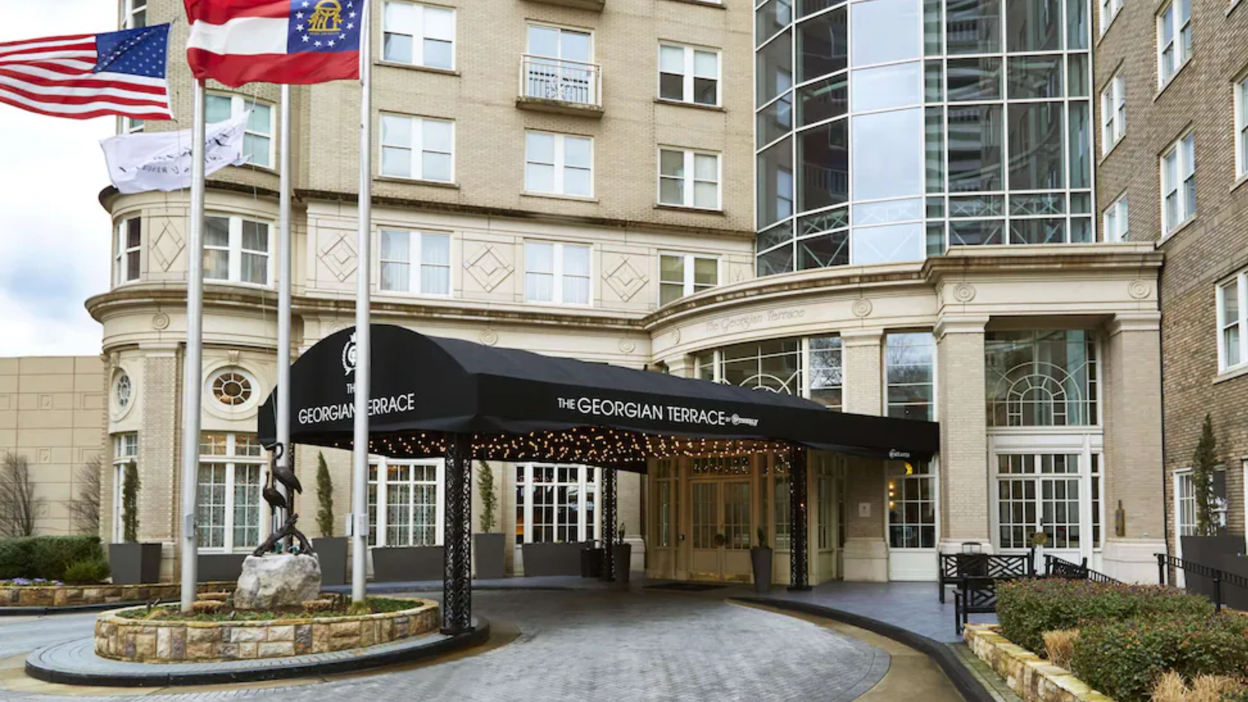 The entrance to The Georgian Terrace hotel featuring a black awning with the hotel's name, surrounded by a circular driveway, flagpoles with flags, and landscaped areas with bushes and flowers.