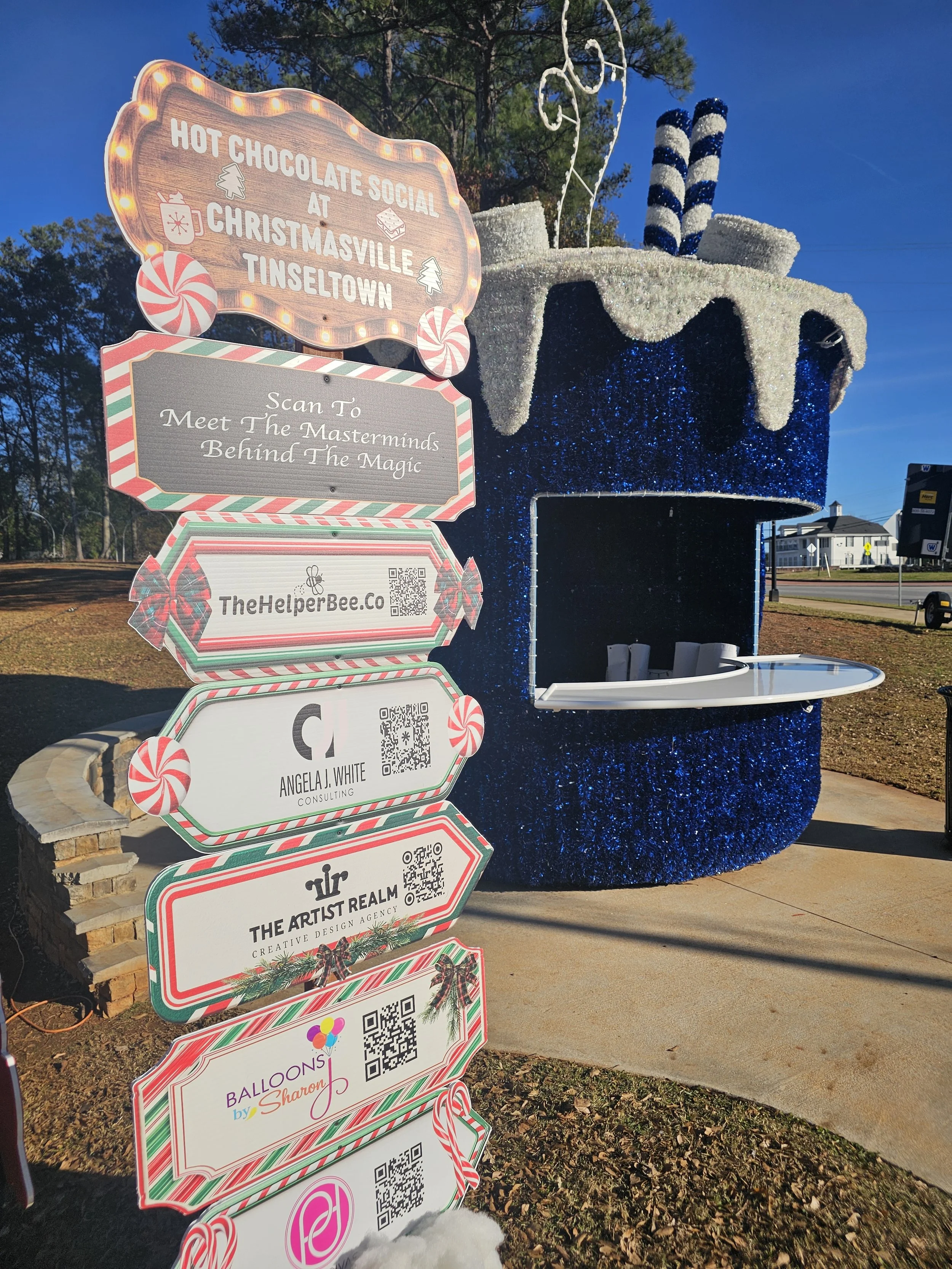 Large holiday-themed booth shaped like a giant cup of hot chocolate with white and blue decorations on top and two candy cane striped poles. In front, a sign with multiple panels invites people to a hot chocolate social at Christmasville Tinseltown, 