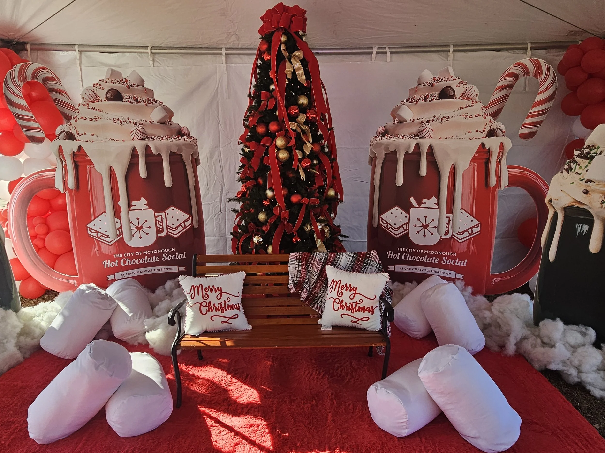 Christmas-themed display featuring two large hot chocolate mugs with whipped cream and candy canes, a decorated Christmas tree in the center, a wooden bench with holiday pillows that read "Merry Christmas," and other festive decorations including red